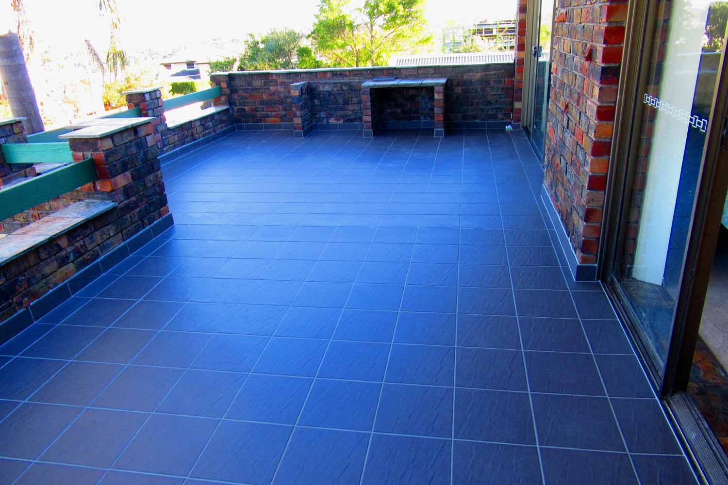 Balcony with newly installed dark gray floor tiles and brick walls with green wooden railing.