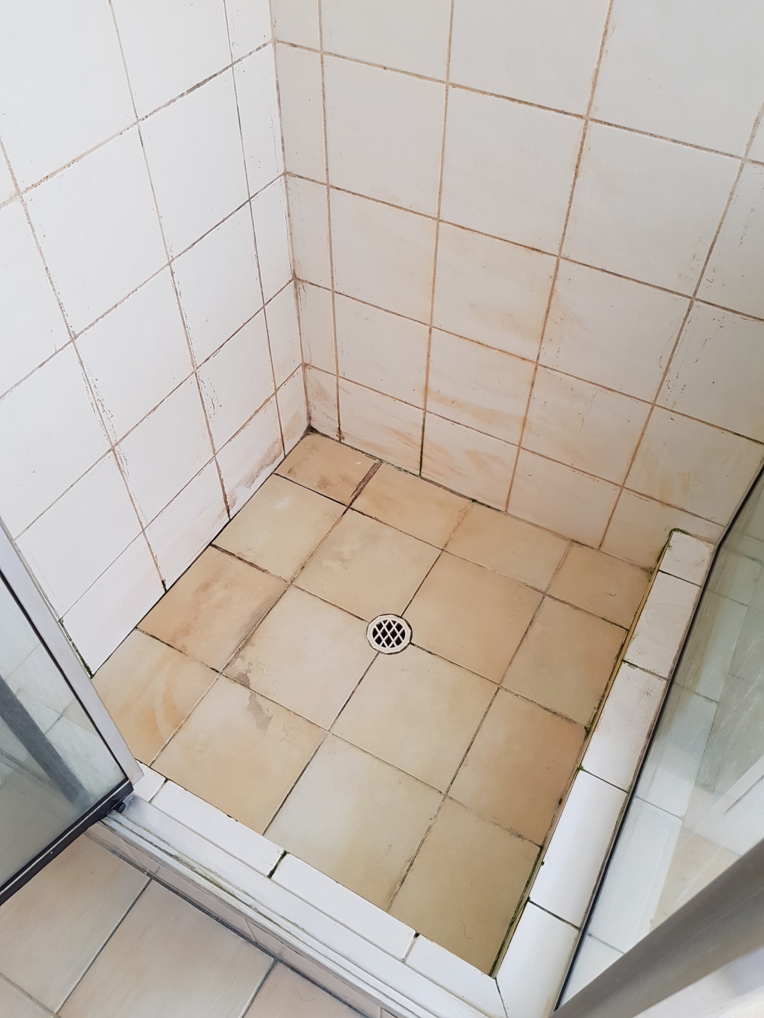 Small square shower stall with beige floor tiles and white wall tiles stained with dirt and mold.