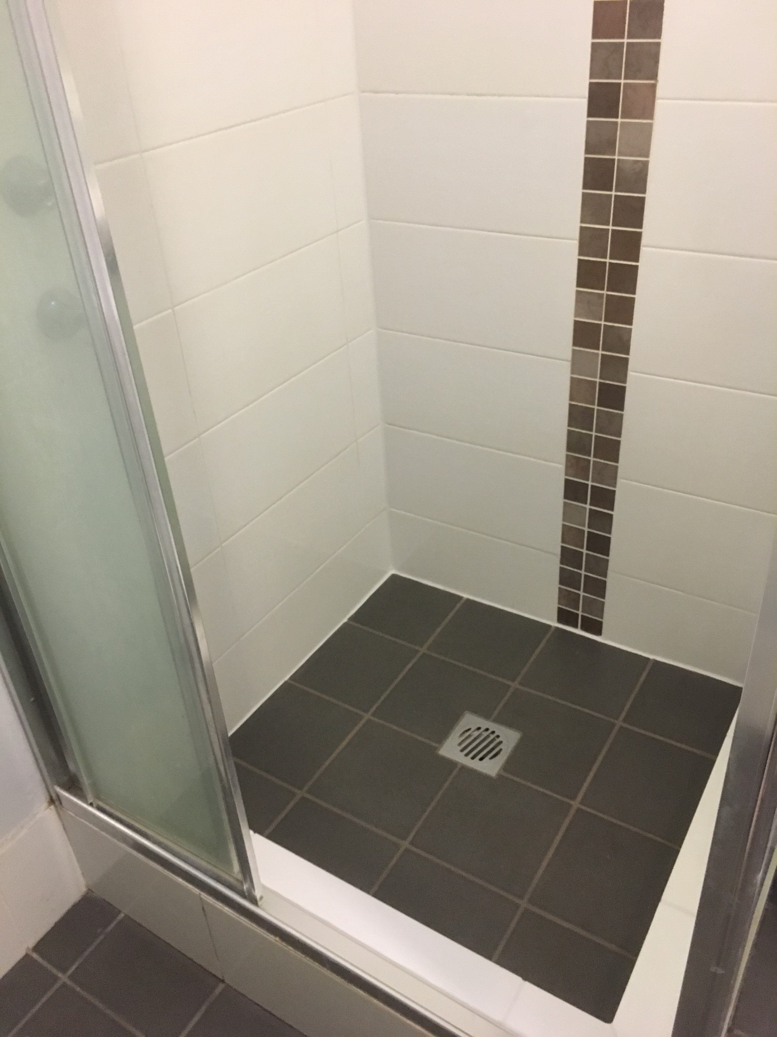 Shower with frosted glass sliding door, dark gray tile floor, and white tiled walls featuring a vertical strip of small brown mosaic tiles.