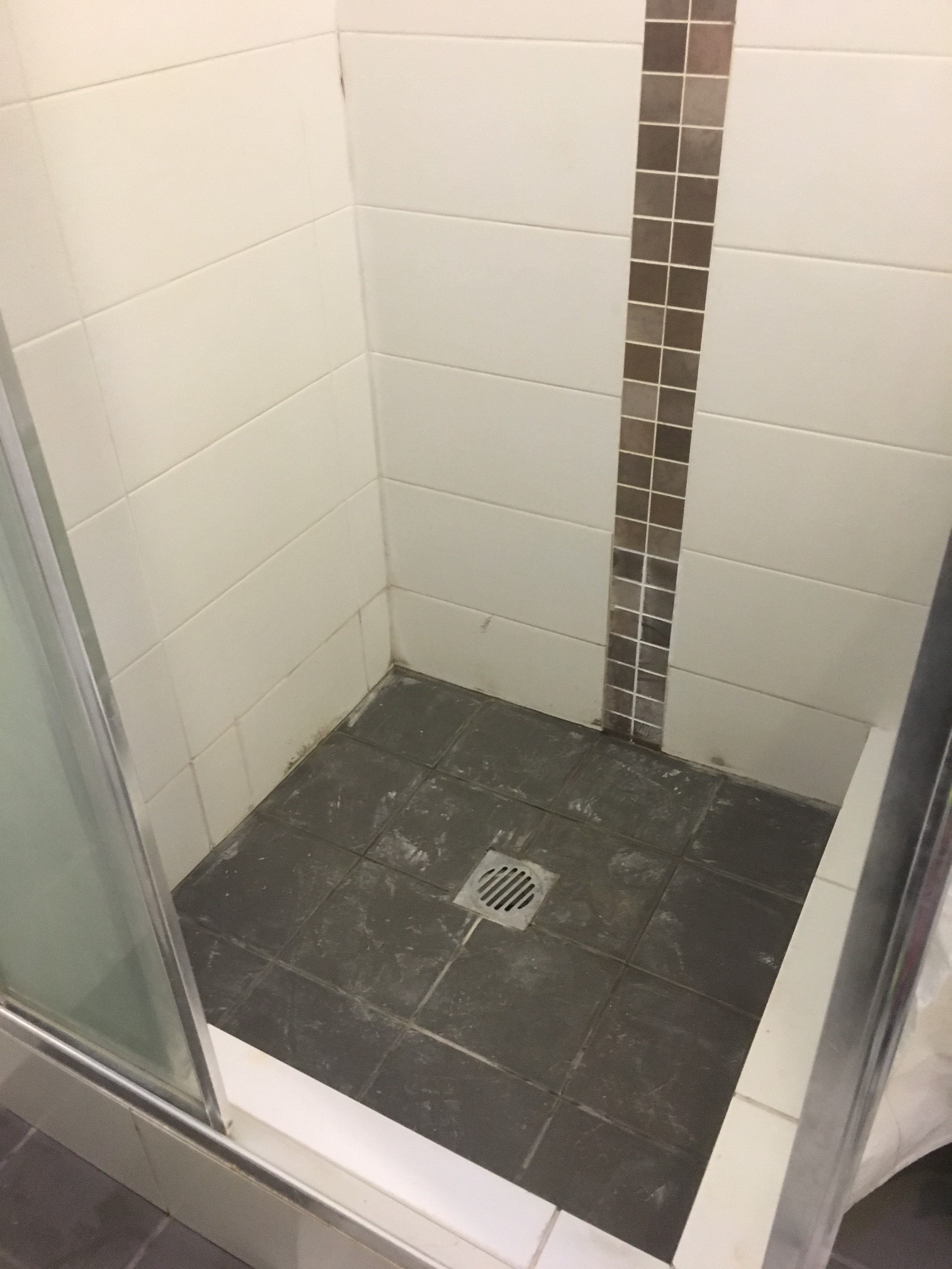 Corner shower stall with white wall tiles, a vertical strip of small brown tiles, and black floor tiles with a central square drain.