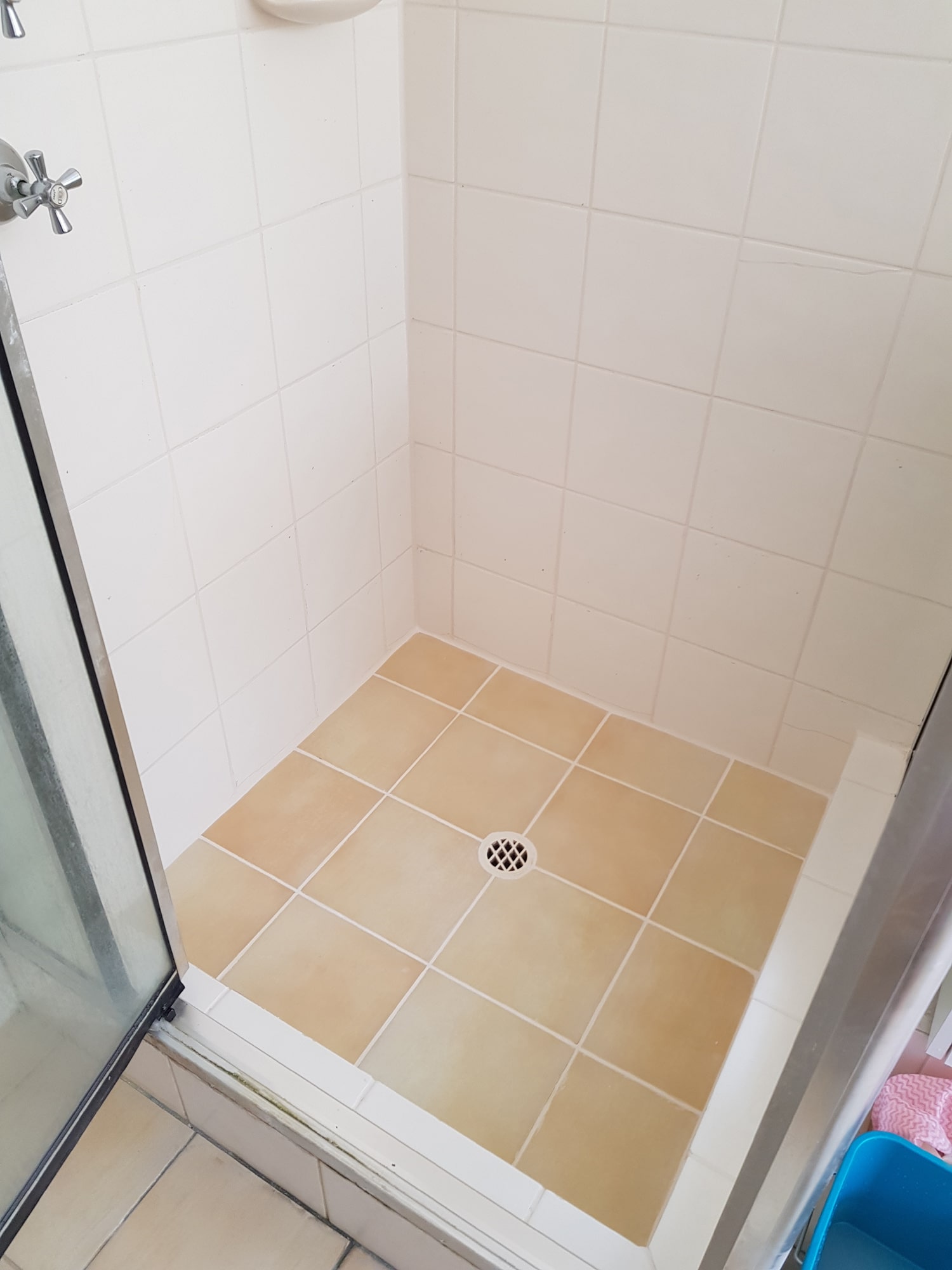 Corner shower area with beige floor tiles and white wall tiles, featuring a central drain and a partially visible sliding glass door.