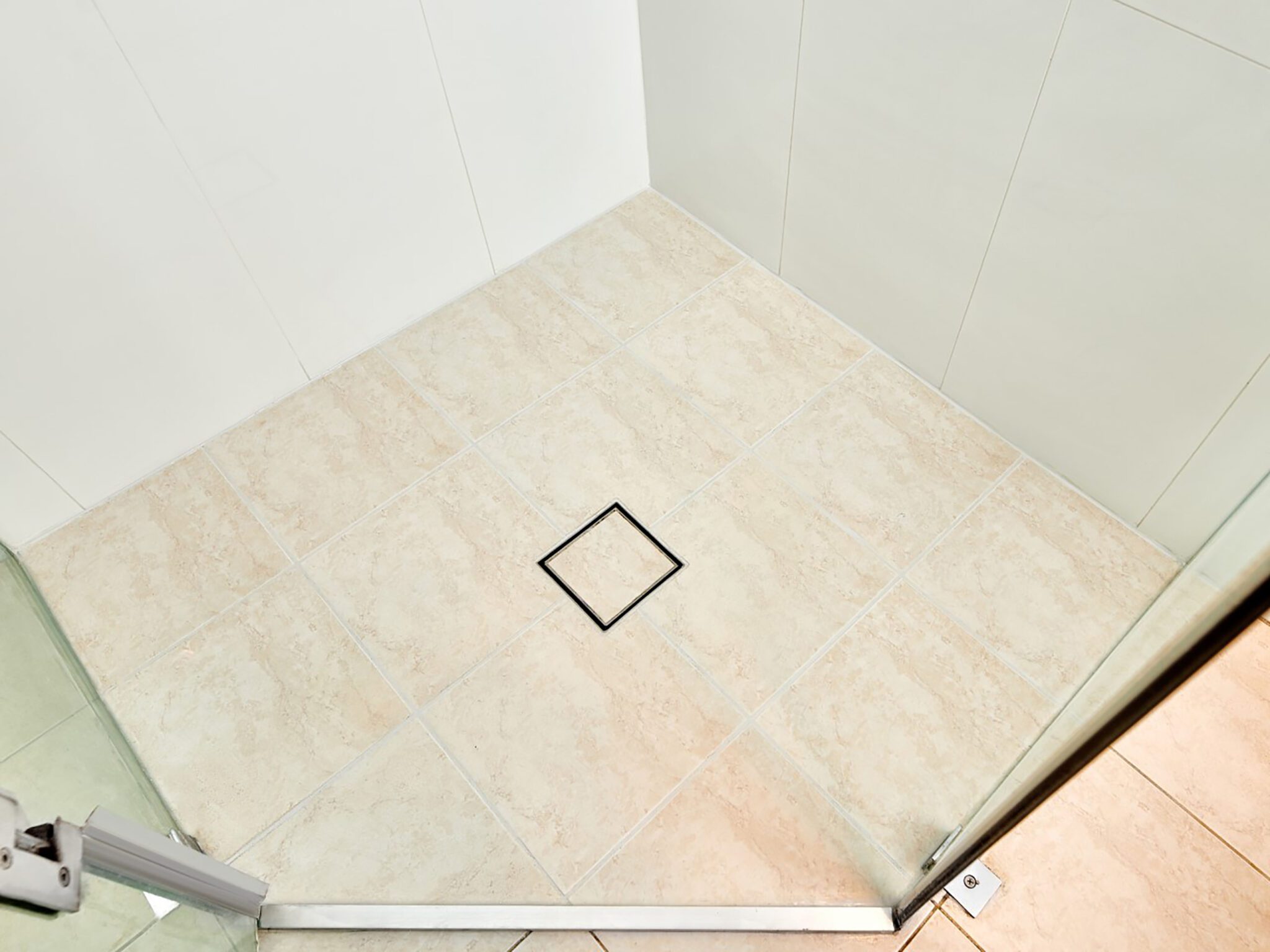 Corner of a tiled shower floor with beige tiles and a square drain in the center, enclosed by glass doors.