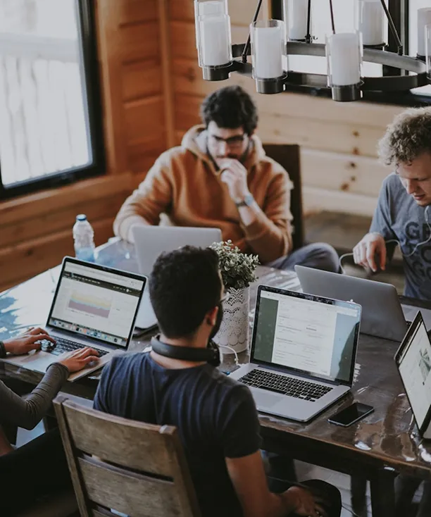Four people working on laptops around a wooden table in a cozy room with wooden walls and a chandelier above.