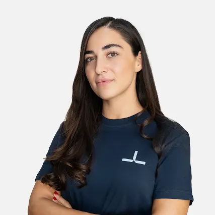 Confident woman with long dark hair wearing a navy blue shirt with a white logo, standing with arms crossed against a plain background.