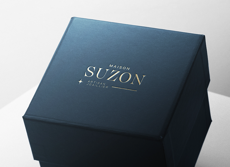 Boîte carrée bleu marine avec le texte doré 'Maison Suzon Artisan Joaillier' sur le dessus.