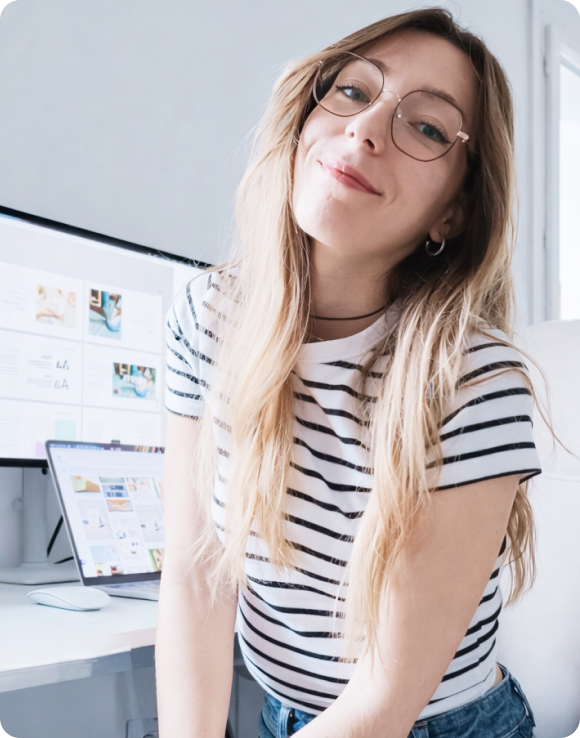 Jeune femme souriante portant des lunettes et un t-shirt rayé blanc et noir, assise près d'un bureau avec un ordinateur et un écran affichant des maquettes de site web.