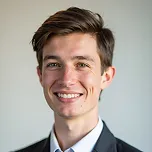 Portrait of a young man with brown hair, wearing a dark suit and white shirt, smiling against a gray background.