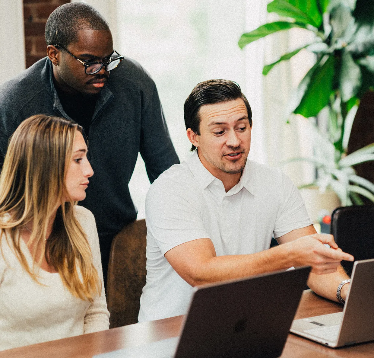 Three colleagues in a meeting room, with one man pointing at a laptop screen while the others listen attentively.