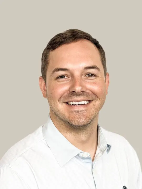Smiling man with short brown hair wearing a white collared shirt against a beige background.