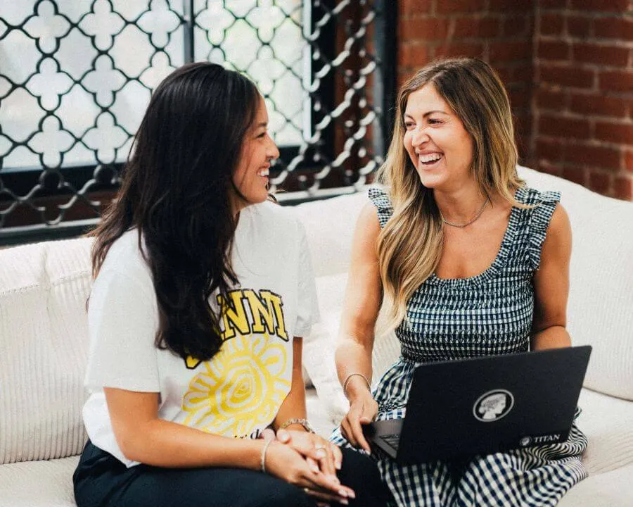 Two women sitting on a white couch, smiling and talking while one holds a laptop with a Titan sticker.