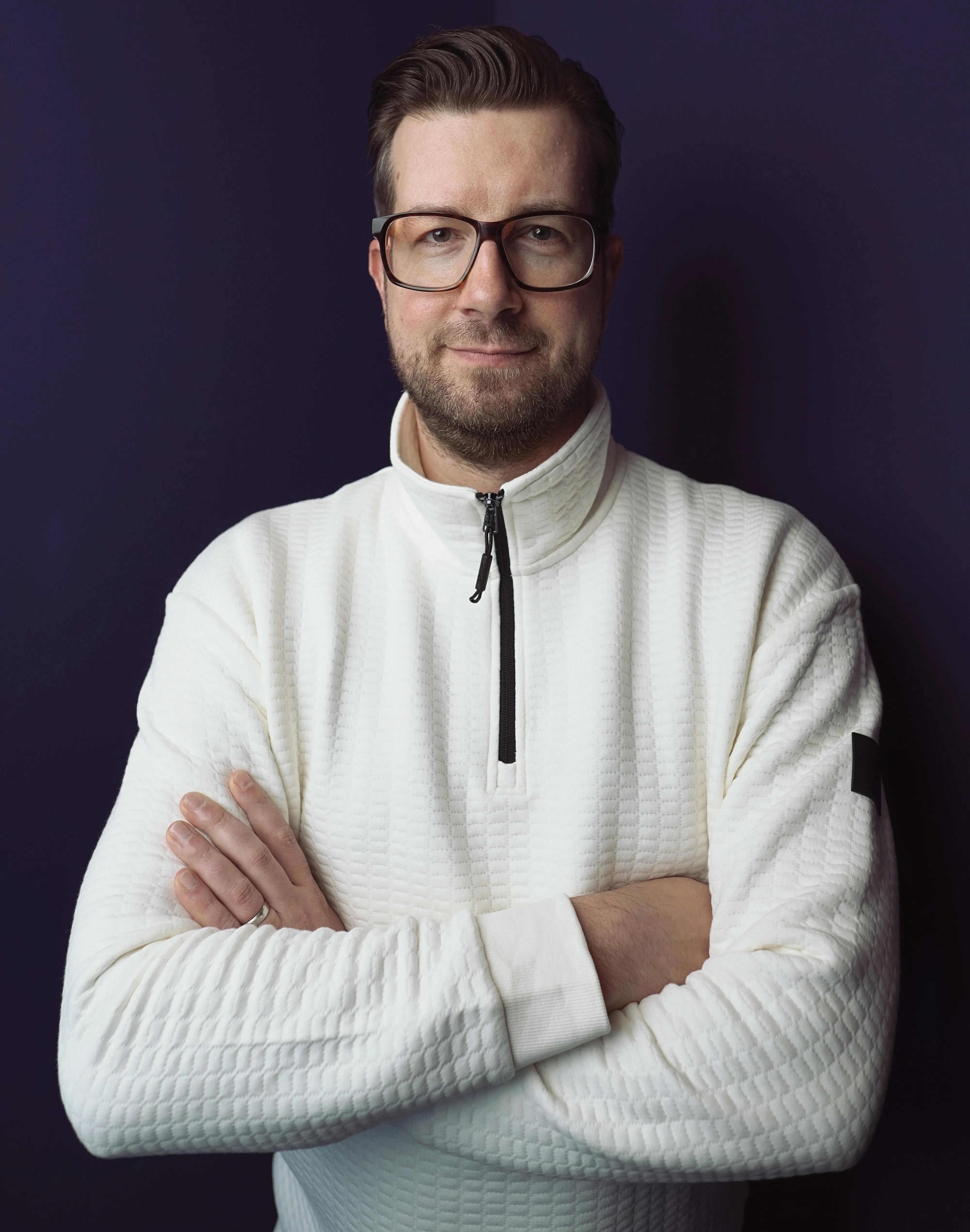 Sascha Albrink trägt einen weißen, strukturierten Pullover und steht mit verschränkten Armen vor einem dunkelvioletten Hintergrund.