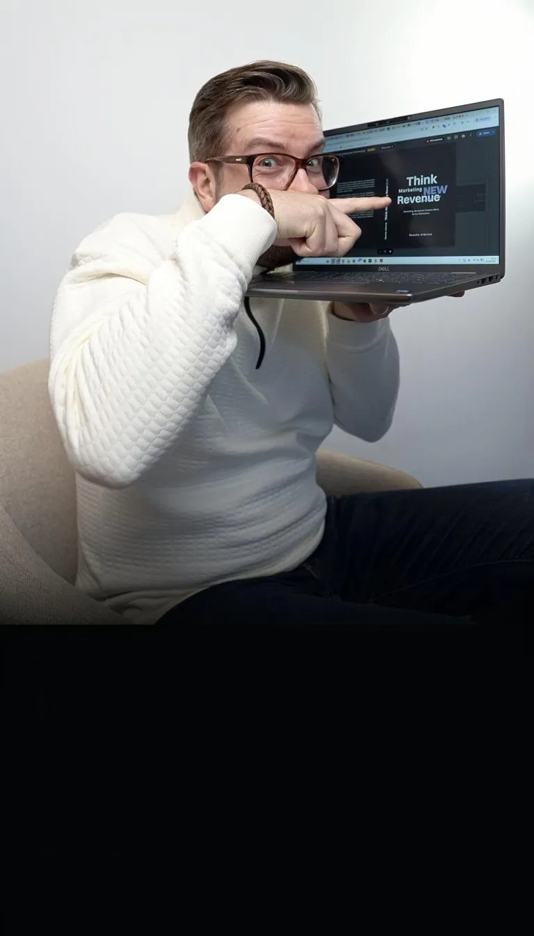 Sascha Albrink mit weißem Pullover sitzt auf einem Stuhl, hält einen Laptop in der Hand und zeigt auf dessen Bildschirm, auf dem ein sein Buch "Think Marketing Revenew" zu sehen ist.