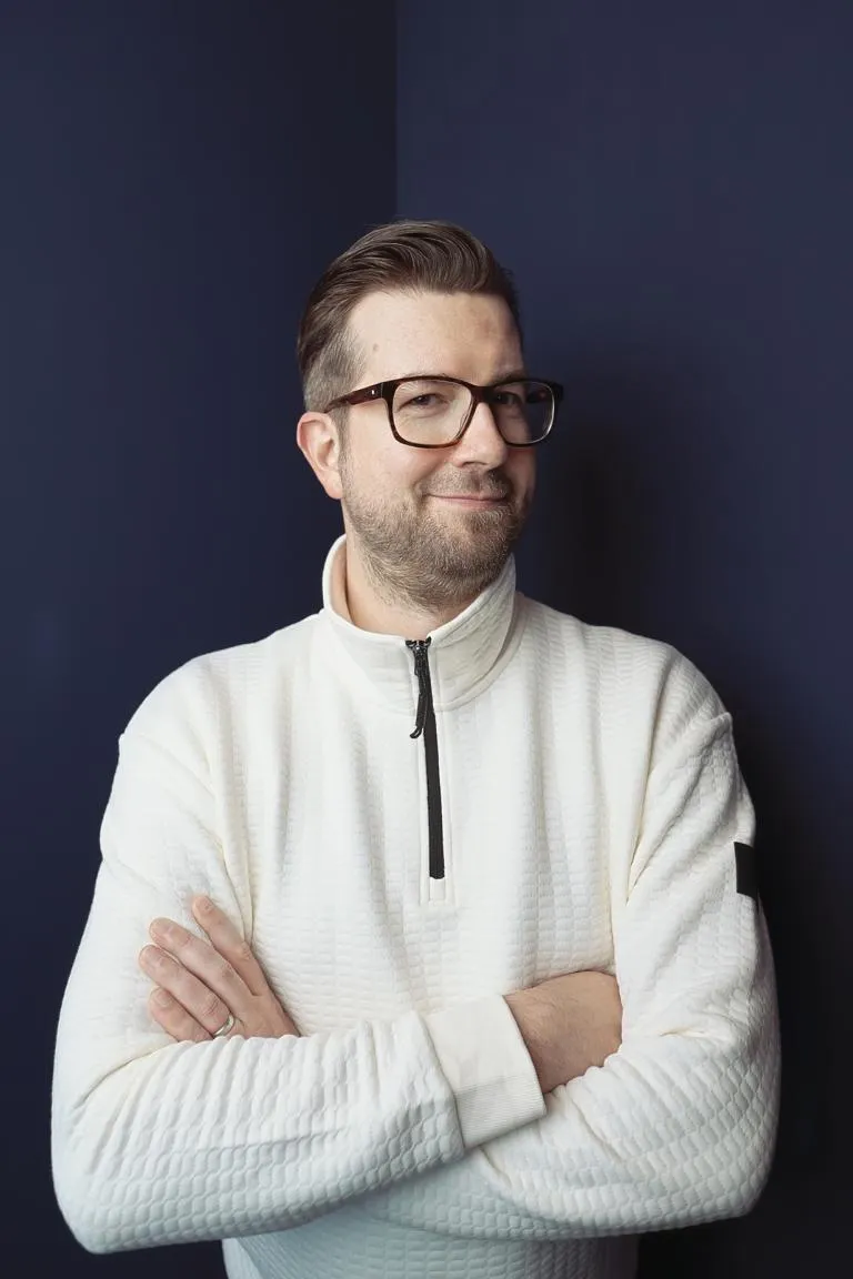 Mann mit kurzen braunen Haaren, Brille und gepflegtem Bart, der einen weißen strukturierten Pullover trägt und die Arme vor einem dunkellila Hintergrund verschränkt hat.