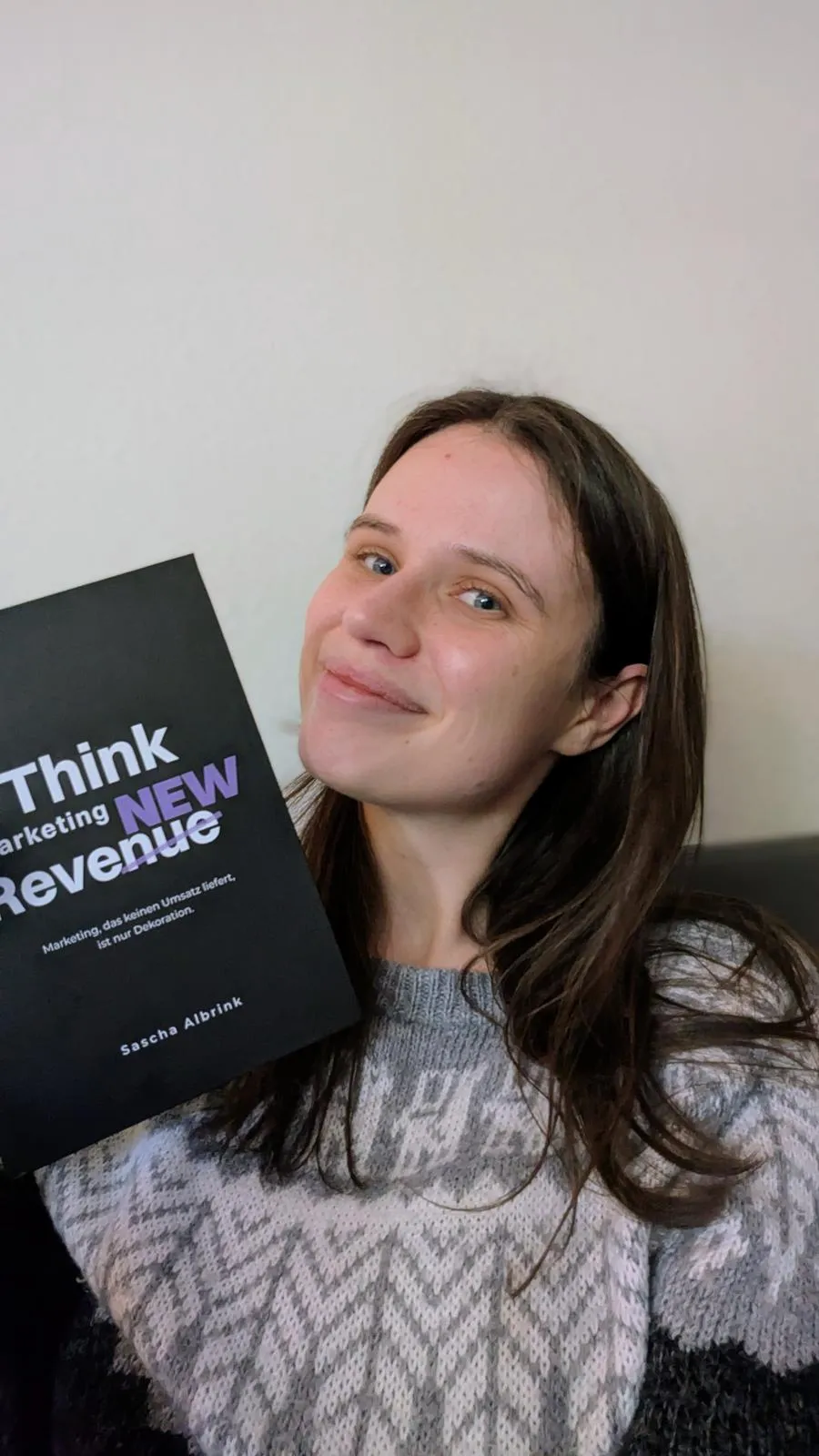Eine Frau in einem gemusterten Pullover, die lächelt und ein Buch mit dem Titel „Think New Revenue“ von Sascha Albrink in der Hand hält.