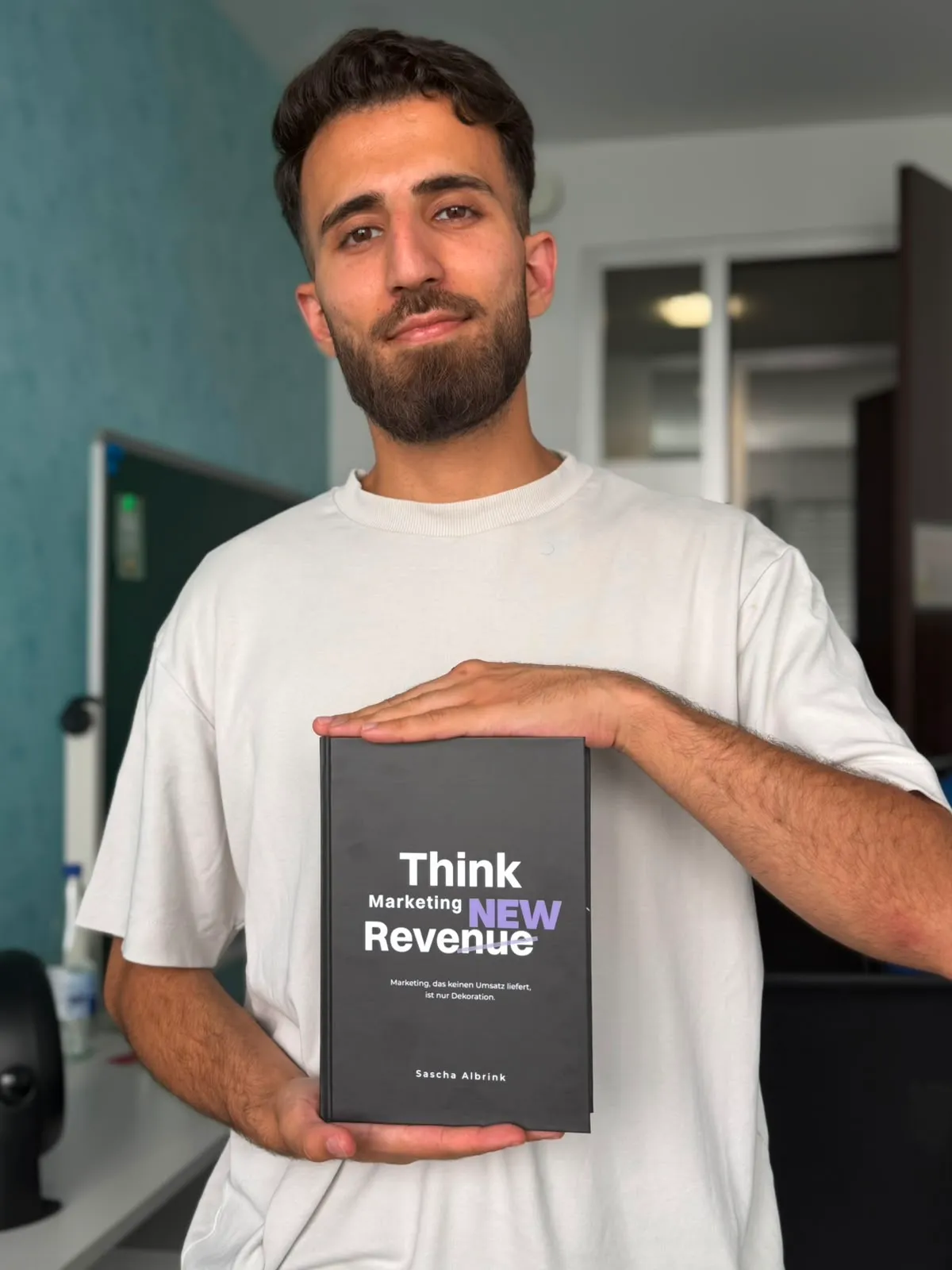 Ein Mann in einem weißen Hemd, der ein Buch mit dem Titel „Think Marketing NEW Revenue“ von Sascha Albrink in der Hand hält.