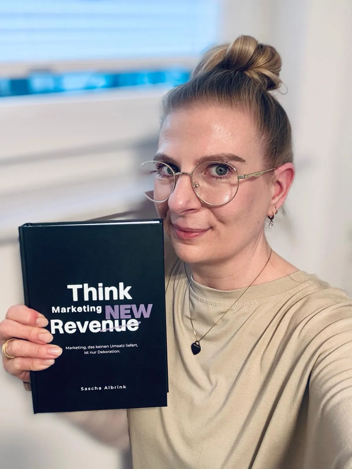 Eine Person mit Brille, die ein Buch mit dem Titel „Think Marketing NEW Revenue“ von Sascha Albrink in der Hand hält.