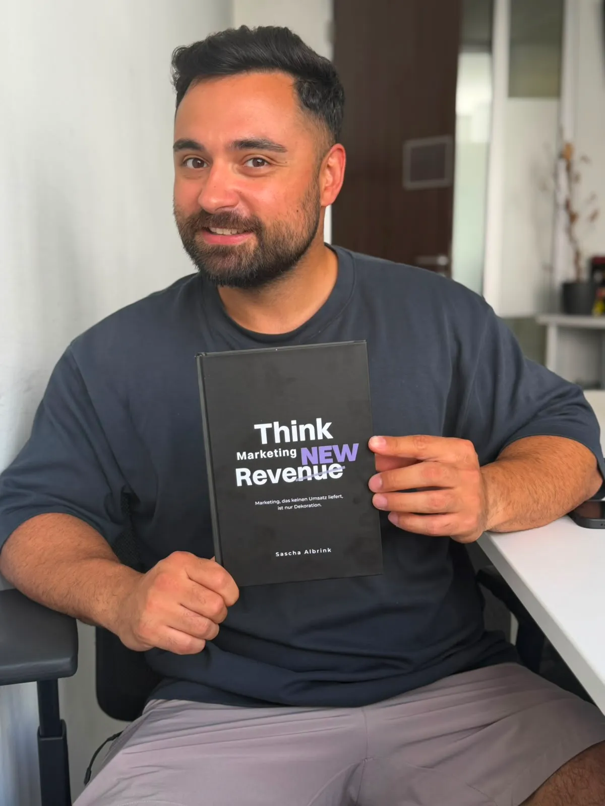 Ein Mann mit Bart, der ein Buch mit dem Titel „Think Marketing NEW Revenue“ von Sascha Albrink in der Hand hält.