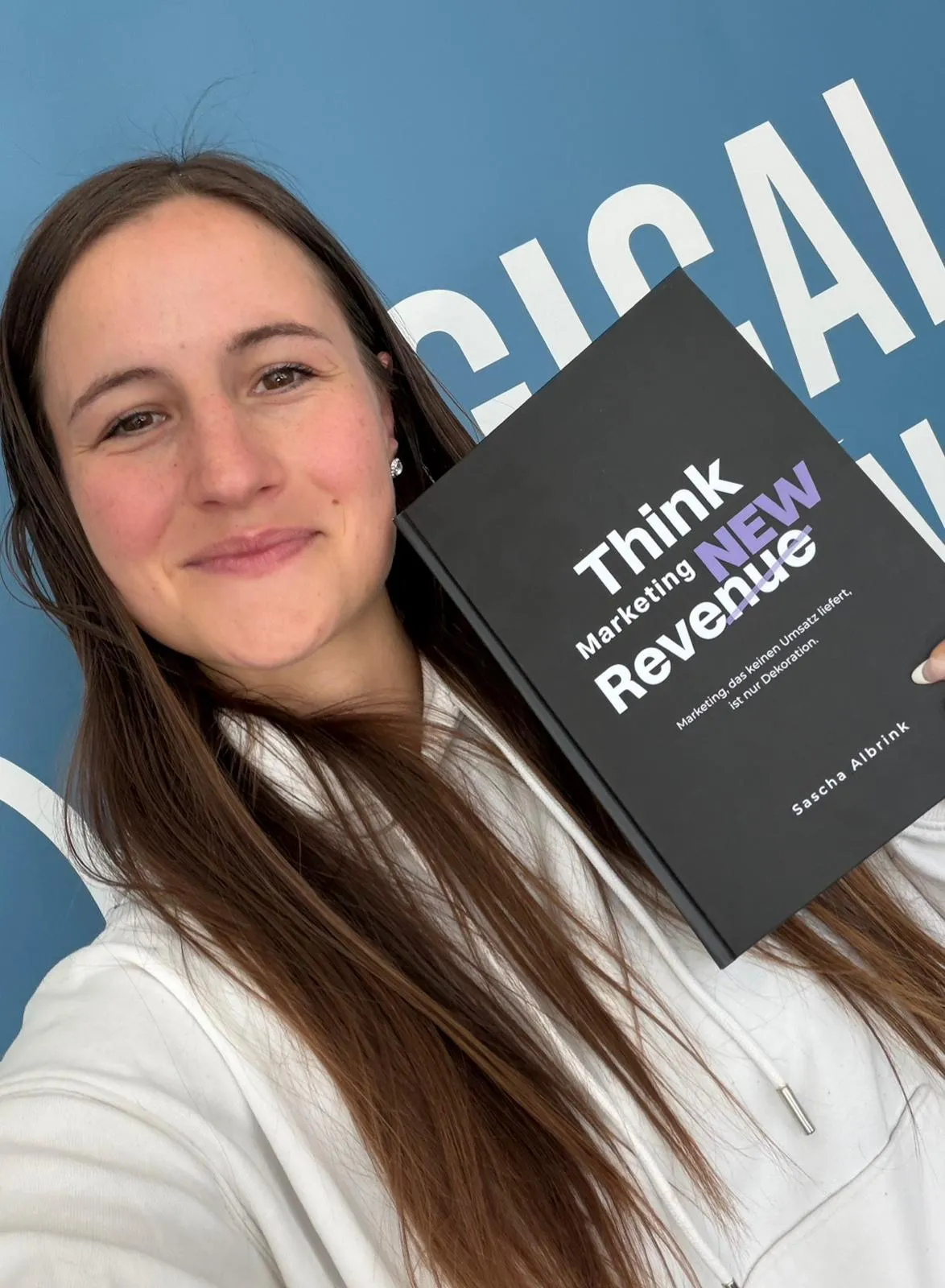 Lächelnde Frau, die vor blauem Hintergrund ein Buch mit dem Titel „Think Marketing New Revenue“ von Sascha Albrink in der Hand hält.