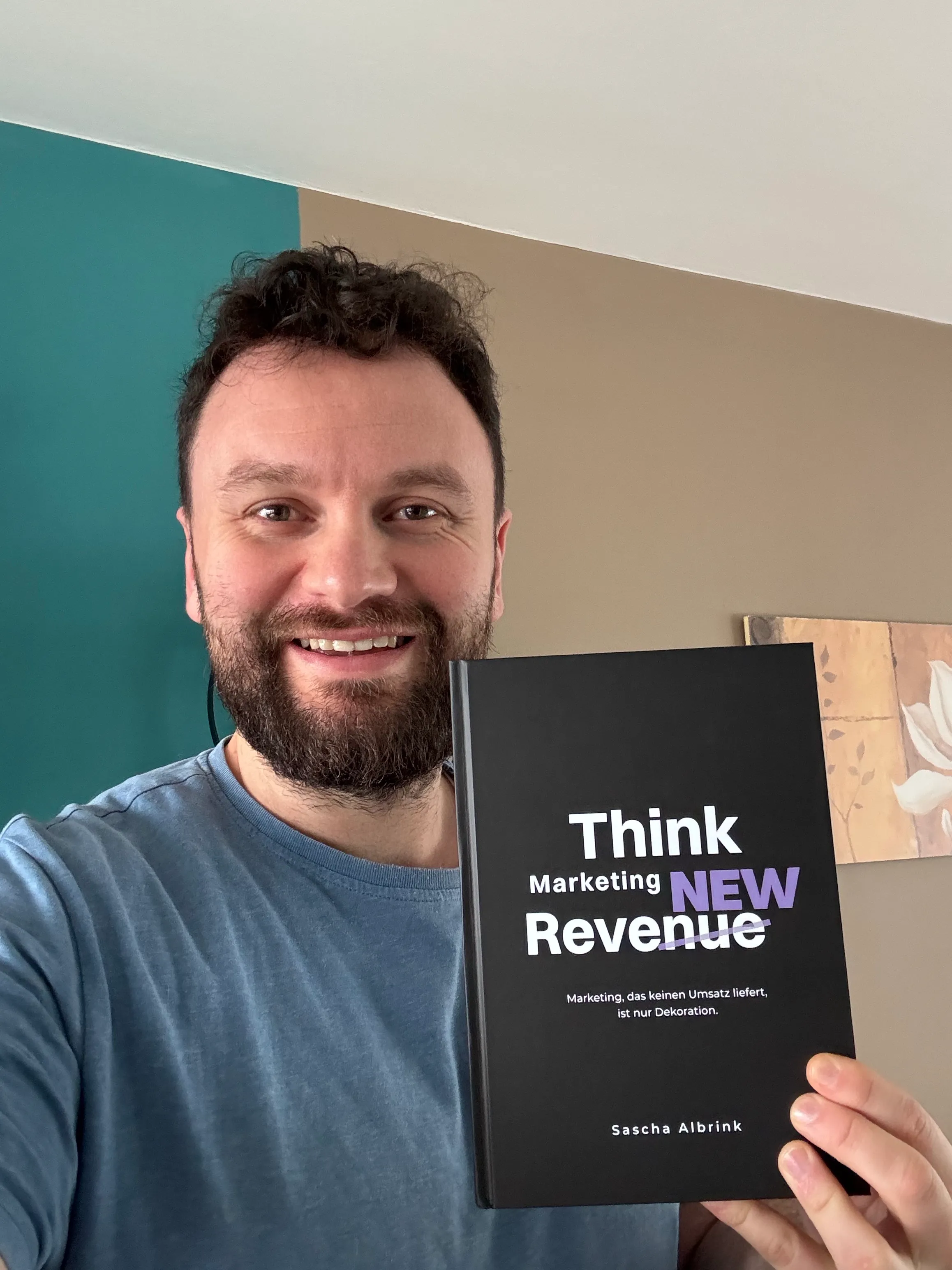 Ein lächelnder Mann mit Bart, der ein schwarzes Buch mit dem Titel „Think Marketing New Revenue“ von Sascha Albrink in der Hand hält.