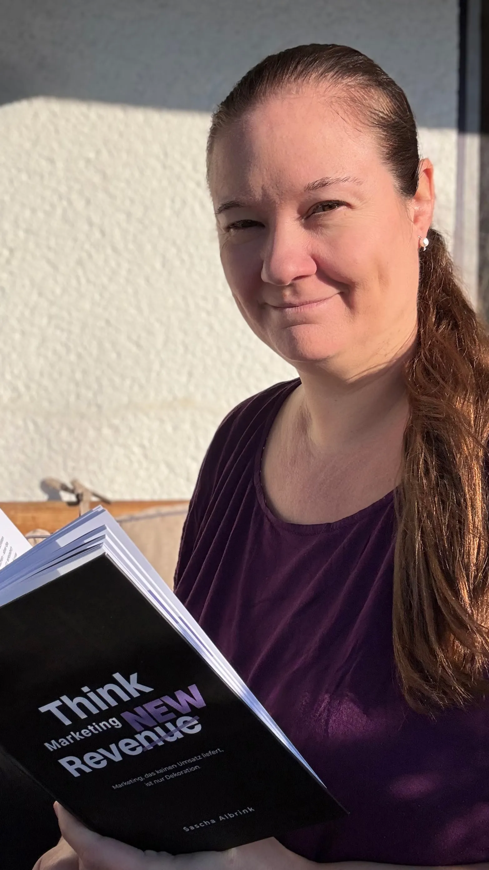 Eine Frau mit langen Haaren, die im Freien im Sonnenlicht ein Buch mit dem Titel „Think Marketing New Revenue“ in der Hand hält.