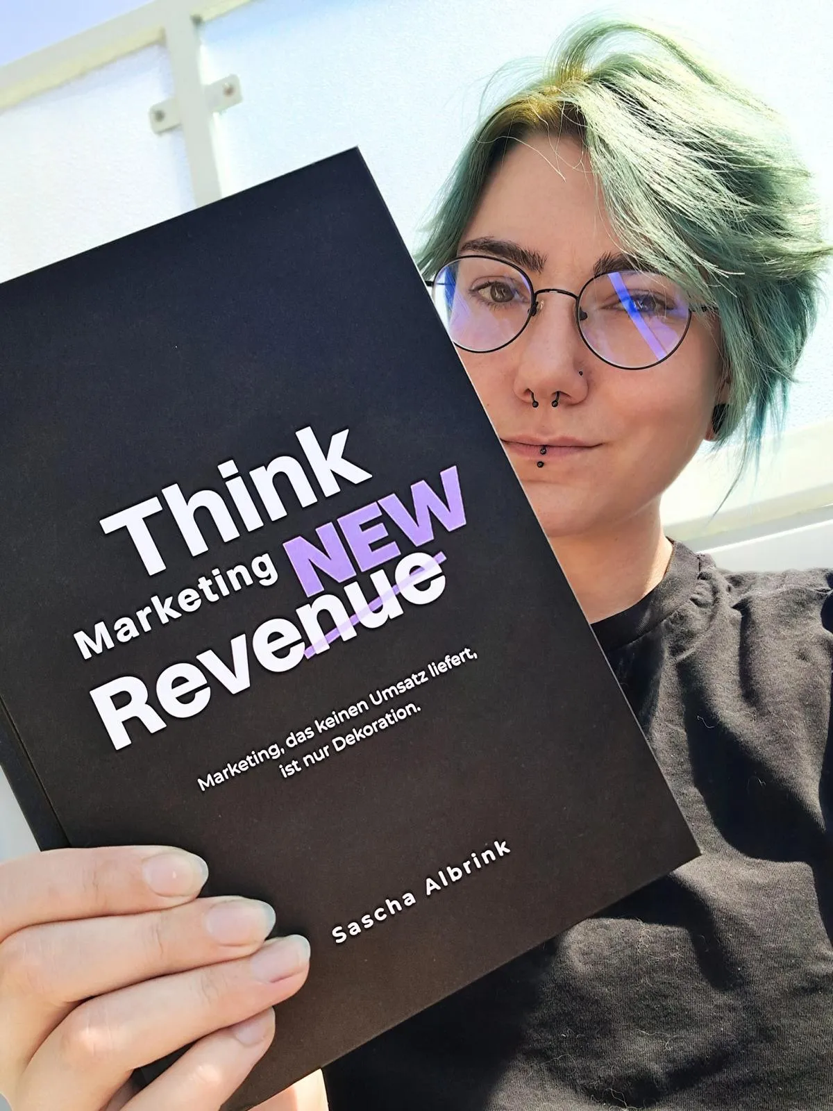 Eine Person mit hellgrünem Haar und Brille, die ein Buch mit dem Titel „Think Marketing NEW Revenue“ von Sascha Albrink in der Hand hält.