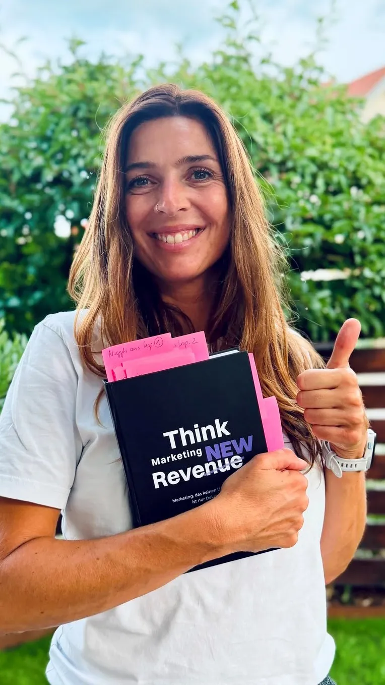 Strahlende Frau hält ein Buch mit dem Titel 'Think Marketing ReveNEW' in der Hand und gibt ein Daumen hoch.