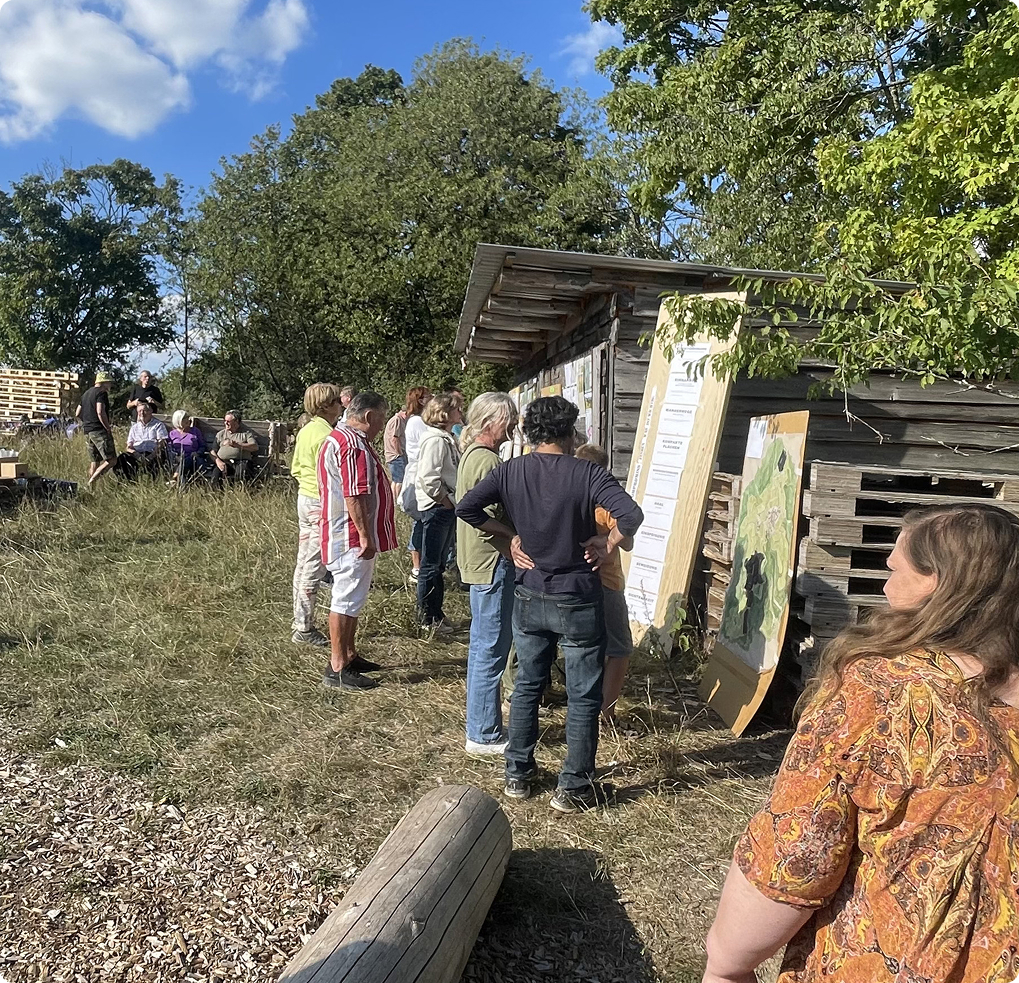 Group of people outdoors gathered around wooden boards with maps or information, near a rustic wooden shed under a clear sky.