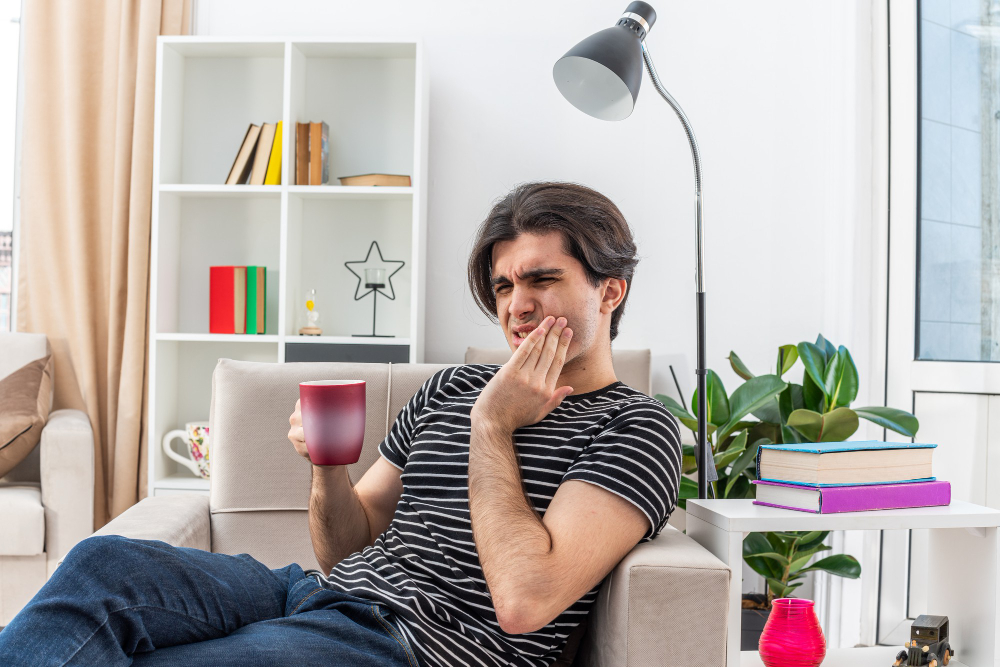 young-man-casual-clothes-holding-cup-hot-tea-looking-unwell-touching-his-cheek-suffering-from-teeth-sensitivity-sitting-chair-light-living-room