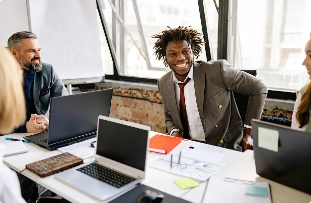 Diverse group of professionals smiling and discussing ideas around a table with laptops and documents in a bright office.