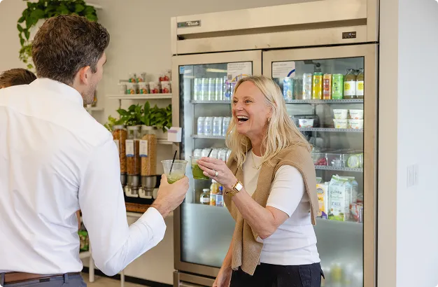 Two people smiling and holding drinks while talking in front of a refrigerated display of beverages and snacks.