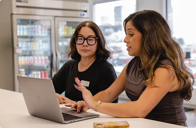 Two women working together at a table with a laptop in a modern office setting with glass-front refrigerators in the background.