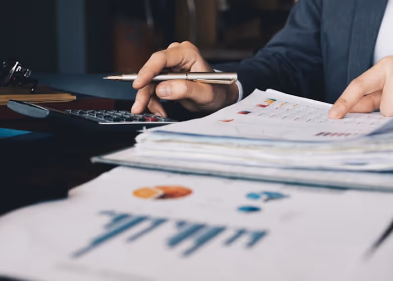 Personne utilisant une calculatrice et tenant un stylo tout en examinant des documents financiers avec des graphiques et des tableaux.