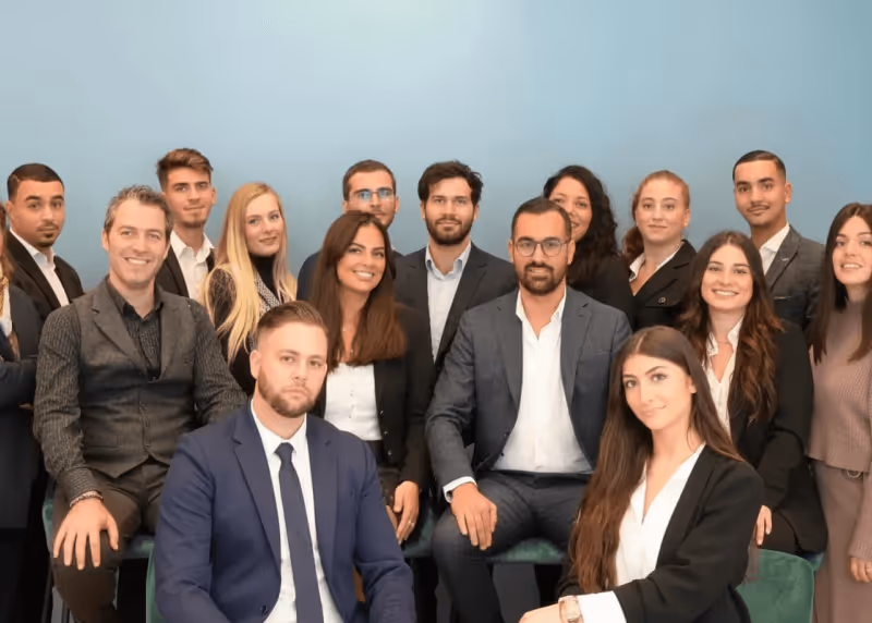 Groupe de 15 professionnels souriants habillés en tenue d'affaires devant un mur bleu clair.