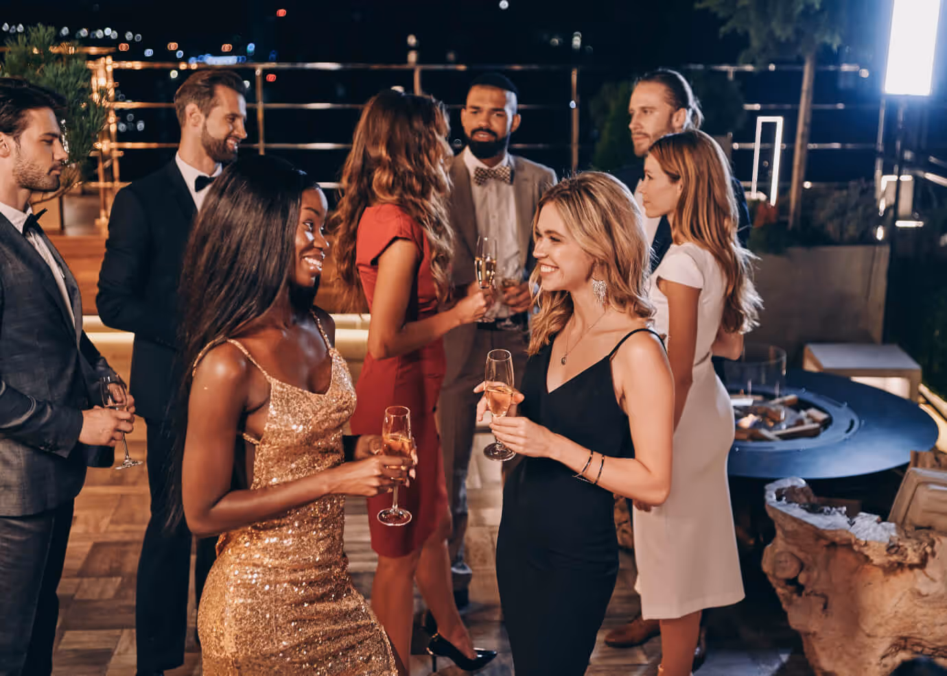 Un groupe de huit jeunes adultes élégamment vêtus discutent et tiennent des flûtes de champagne lors d'une soirée en plein air.