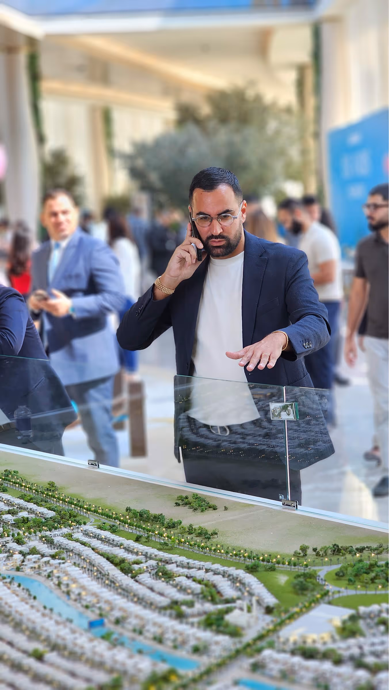 Homme en veste sombre parlant au téléphone en observant une maquette urbaine détaillée dans un espace public animé.