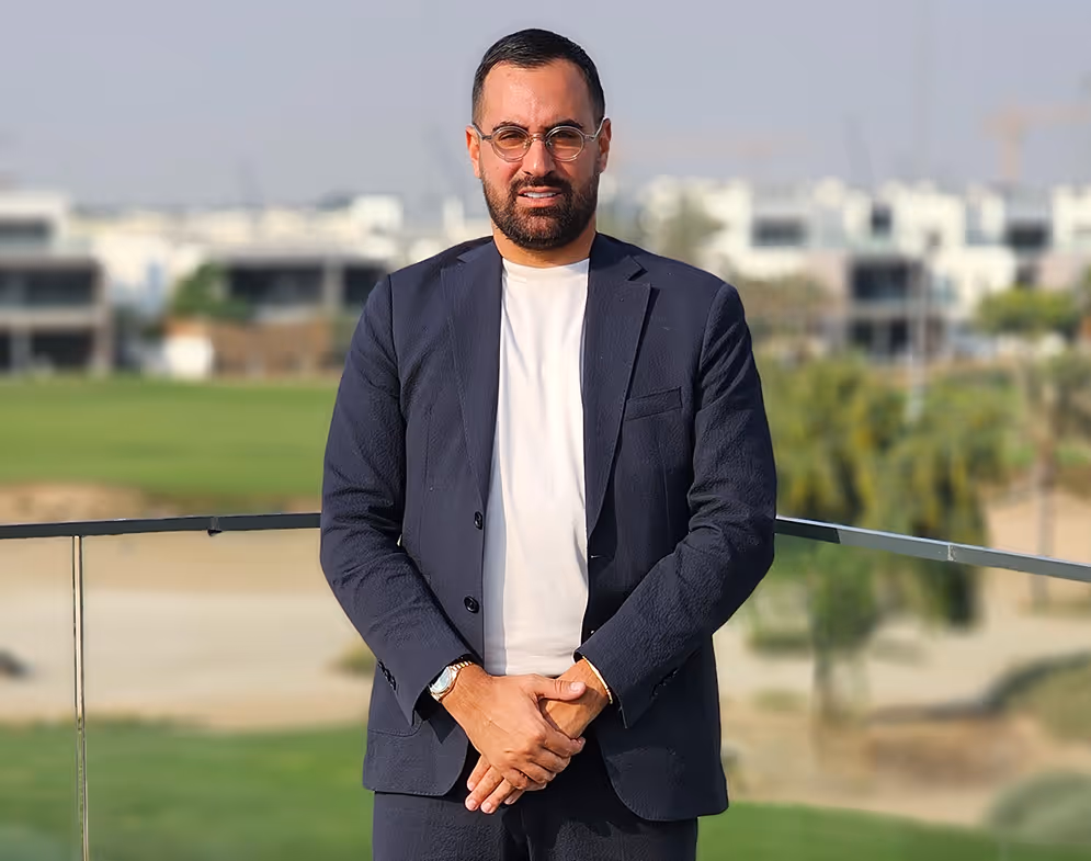Un homme portant des lunettes, un costume bleu foncé et un t-shirt blanc, posant sur un balcon avec un paysage flou d'herbe et de bâtiments en arrière-plan.