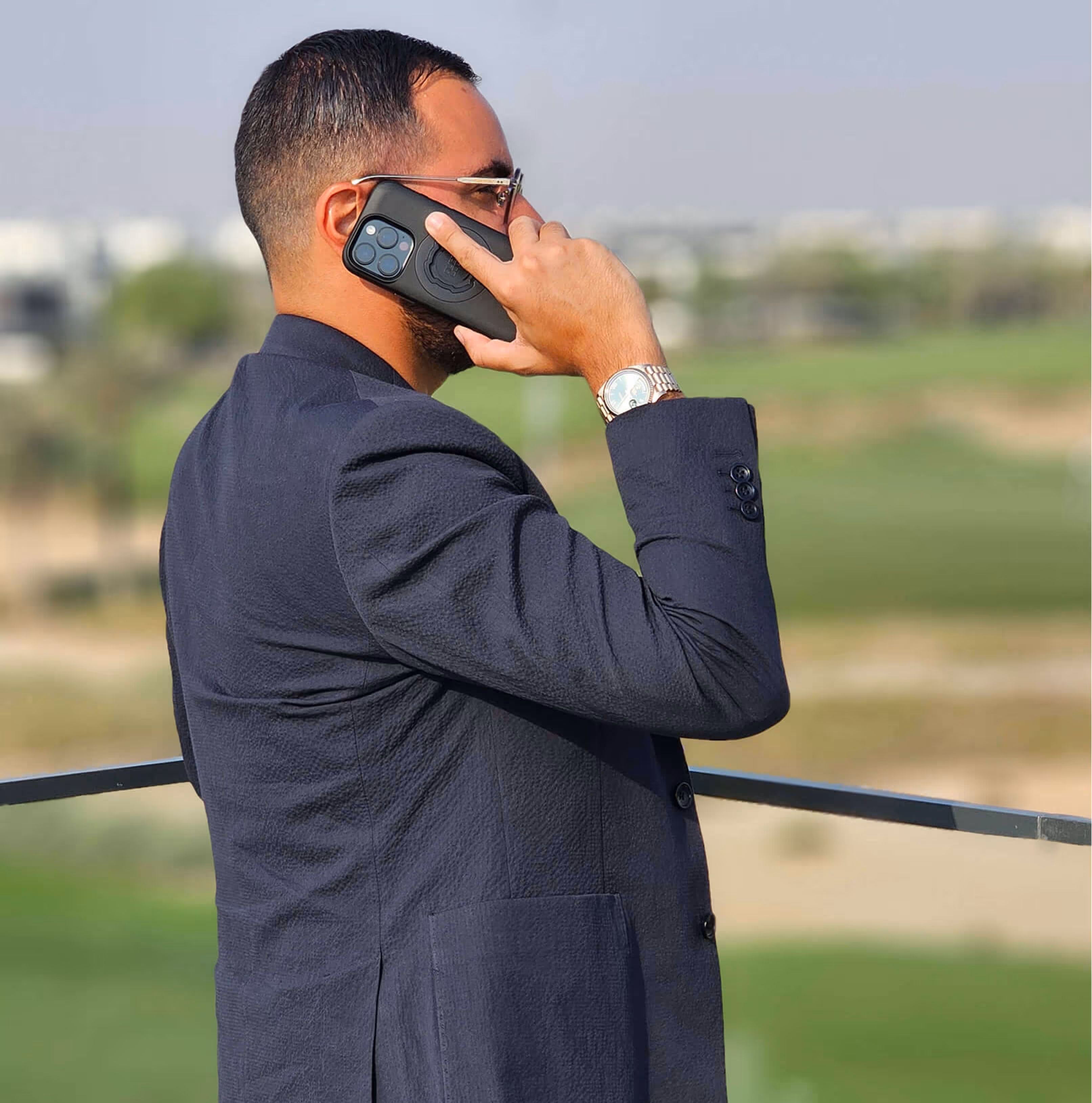 Homme en costume bleu parlant au téléphone à l'extérieur avec un paysage flou en arrière-plan.