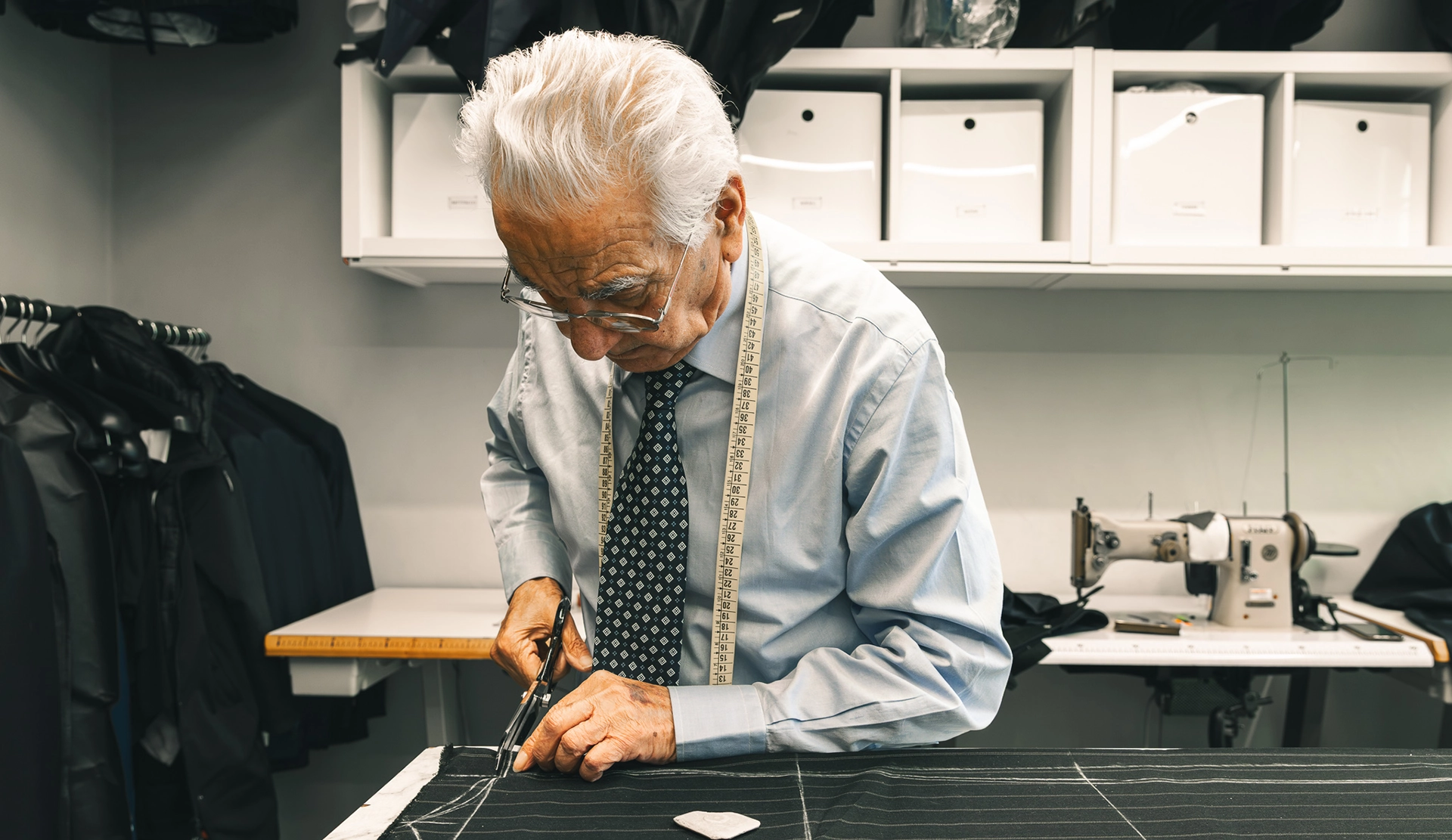 Sarto di Zeolla al lavoro sul tavolo di taglio: artigiano concentrato mentre misura e prepara il tessuto per un abito su misura, immagine che racconta il savoir-faire sartoriale dell’atelier di moda maschile, foto Digistart.
