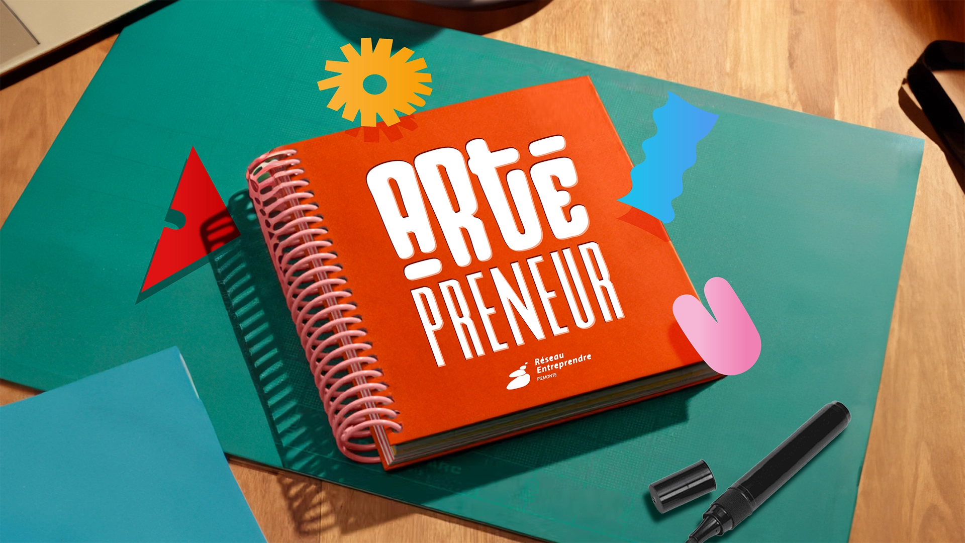 Visual identity per l’evento “Artepreneur” di Réseau Entreprendre Piemonte: taccuino arancione con lettering creativo e elementi grafici colorati su scrivania, parte del design coordinato realizzato da Digistart, agenzia creativa a Torino.