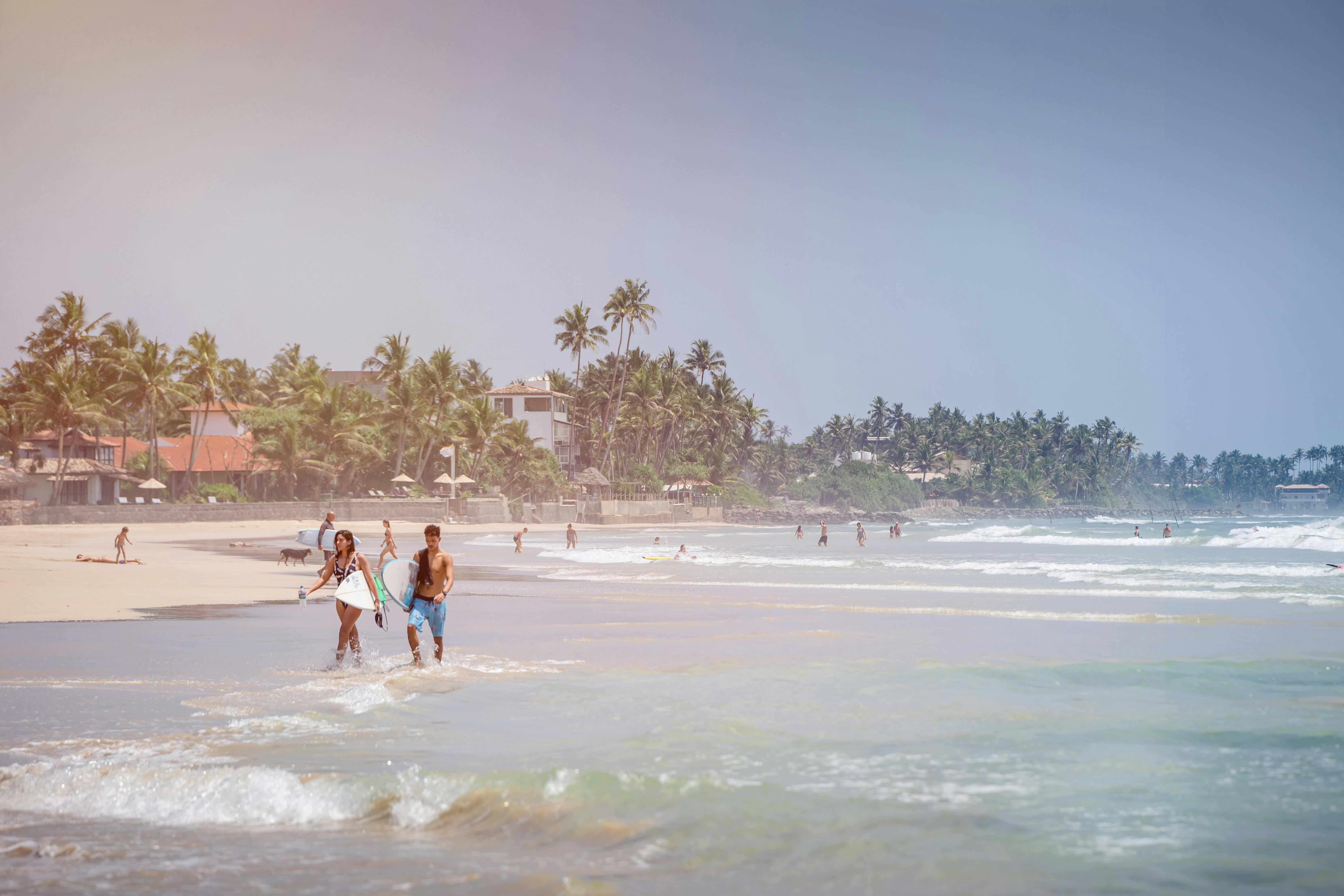 A bright, sun-drenched tropical beach in Sri Lanka featuring two surfers walking along the shoreline with their boards as waves roll in.