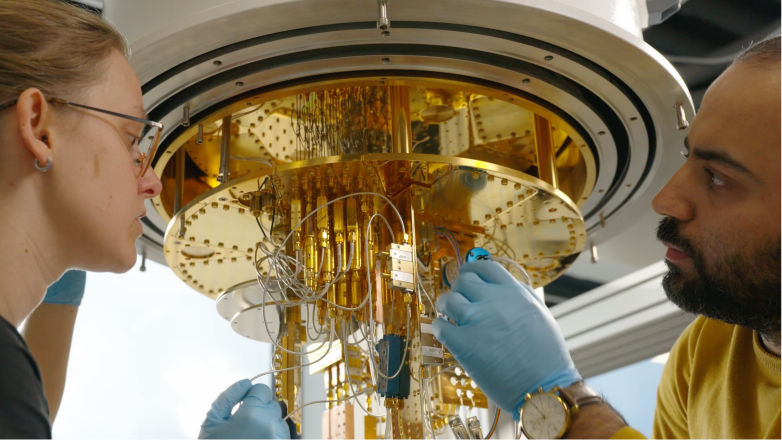 Two scientists wearing blue gloves closely examine and adjust gold-colored components inside a large, circular scientific instrument.