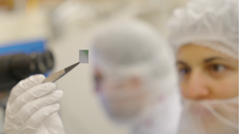 Scientist wearing protective gear holding a small microchip with tweezers in a laboratory.