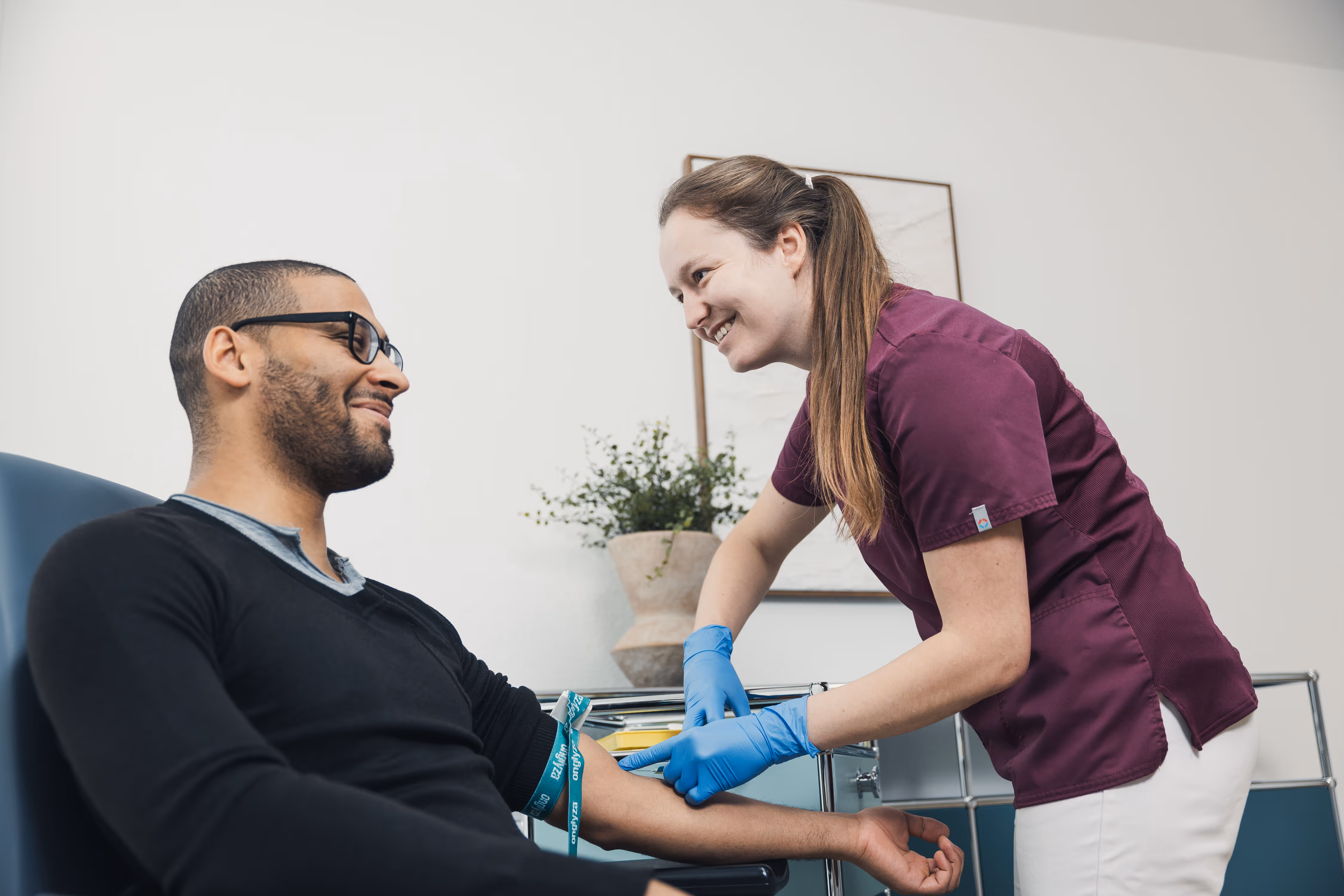 Healthcare worker in purple scrubs smiling while preparing a man's arm for a blood draw.