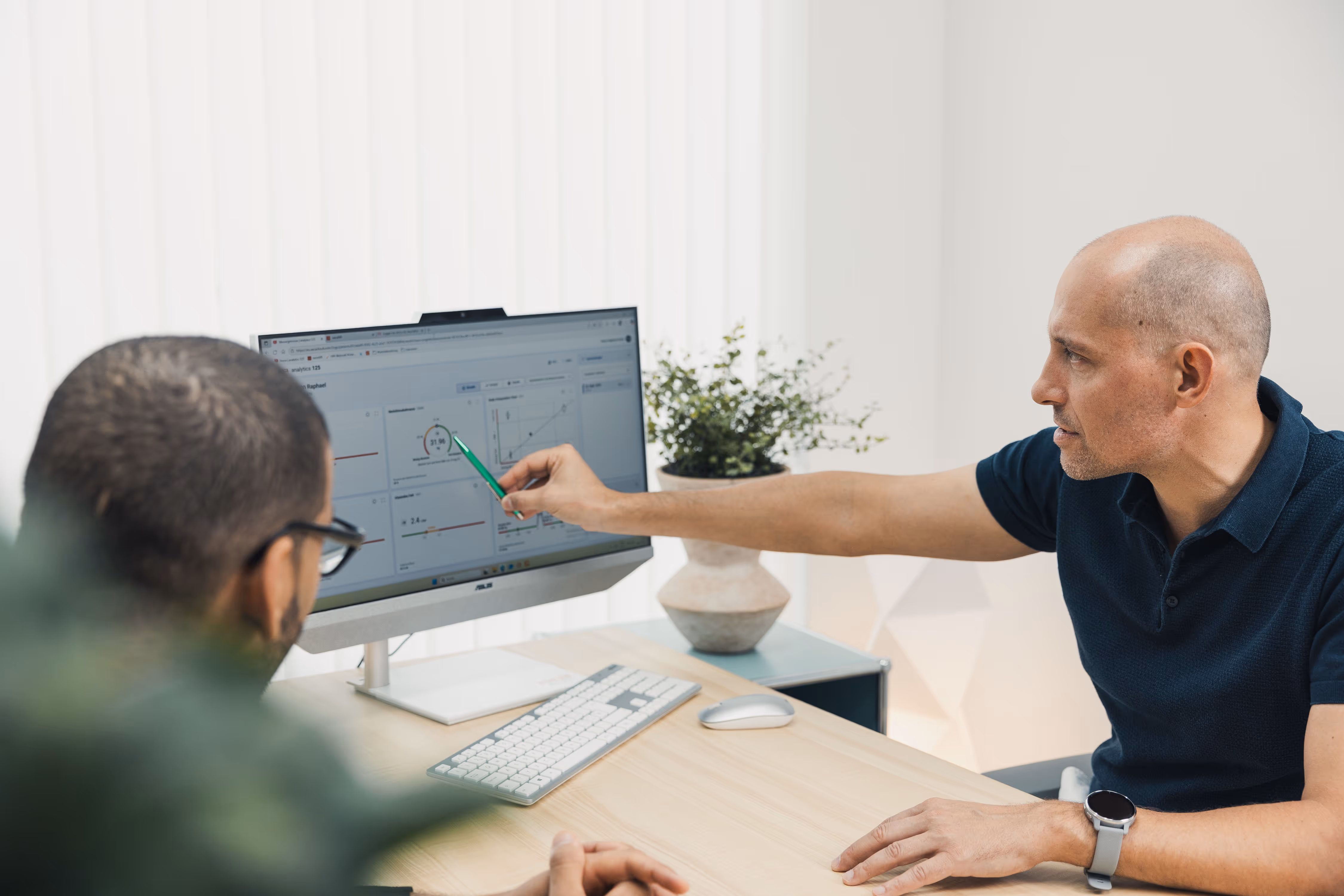 Man pointing with a green pen at data charts on a desktop computer screen while another man watches attentively.