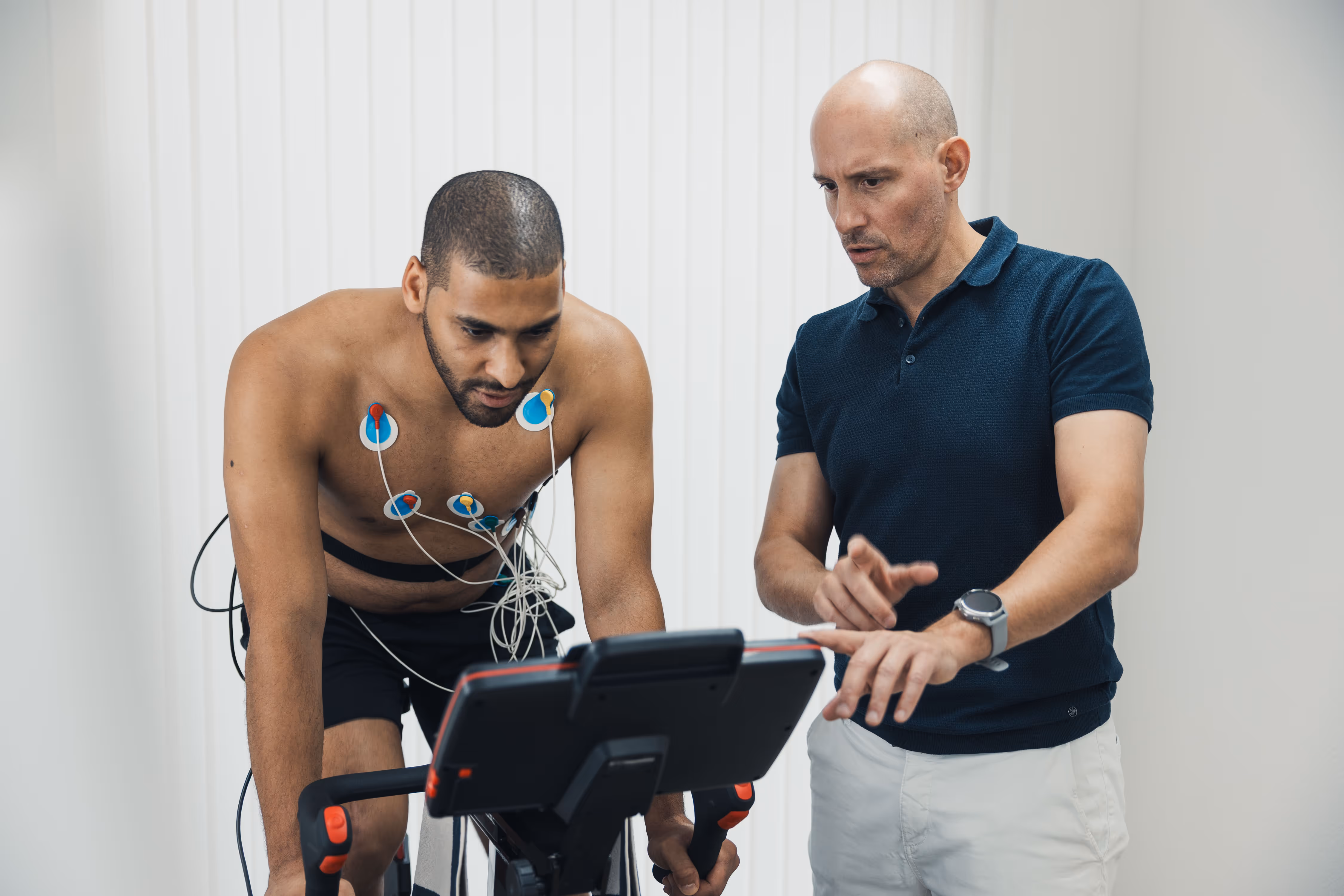Man on stationary bike with ECG sensors attached to his chest, monitored by a trainer pointing at the bike's display.