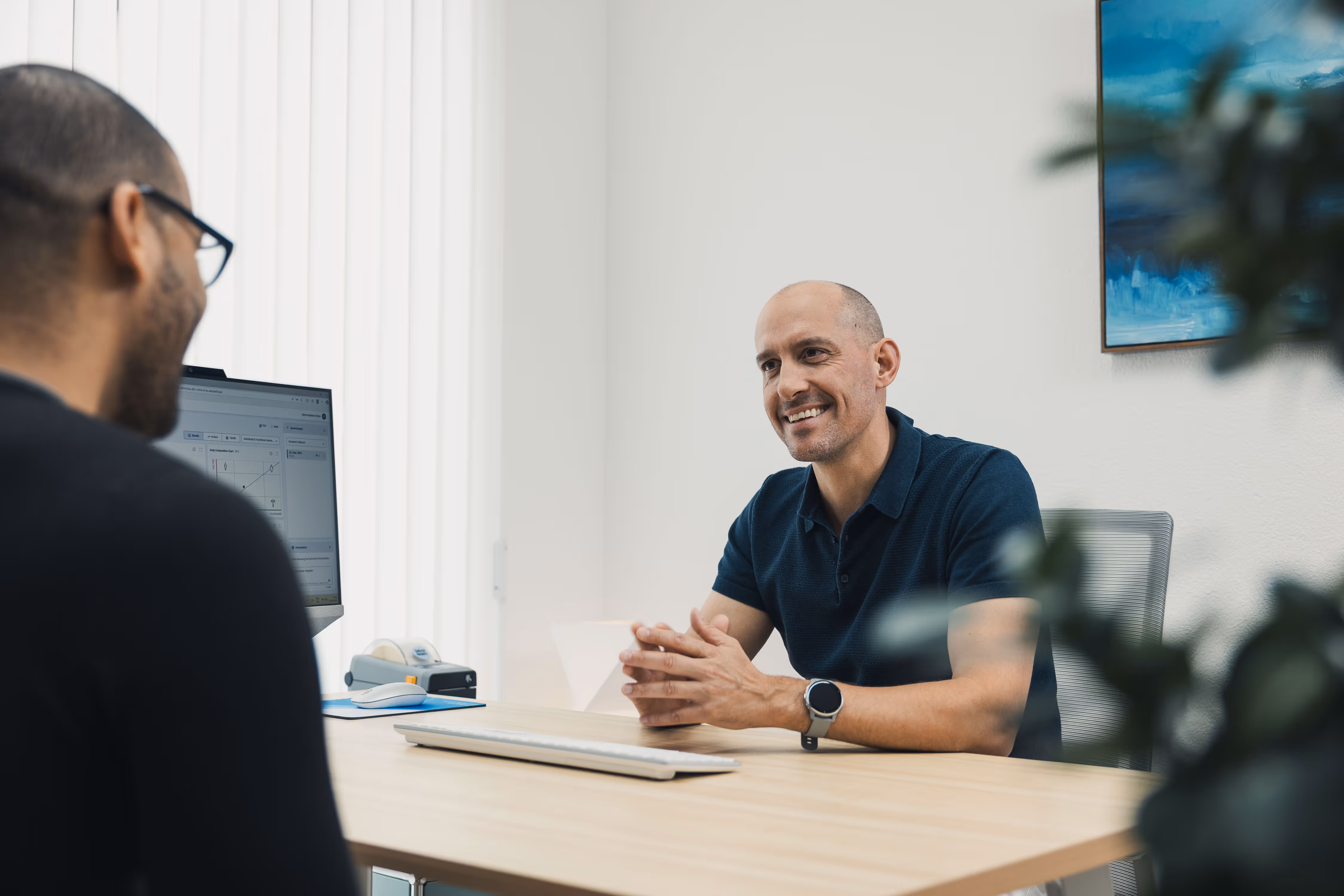 Two men having a conversation across a desk in a bright office, with one smiling and the other facing a computer screen.