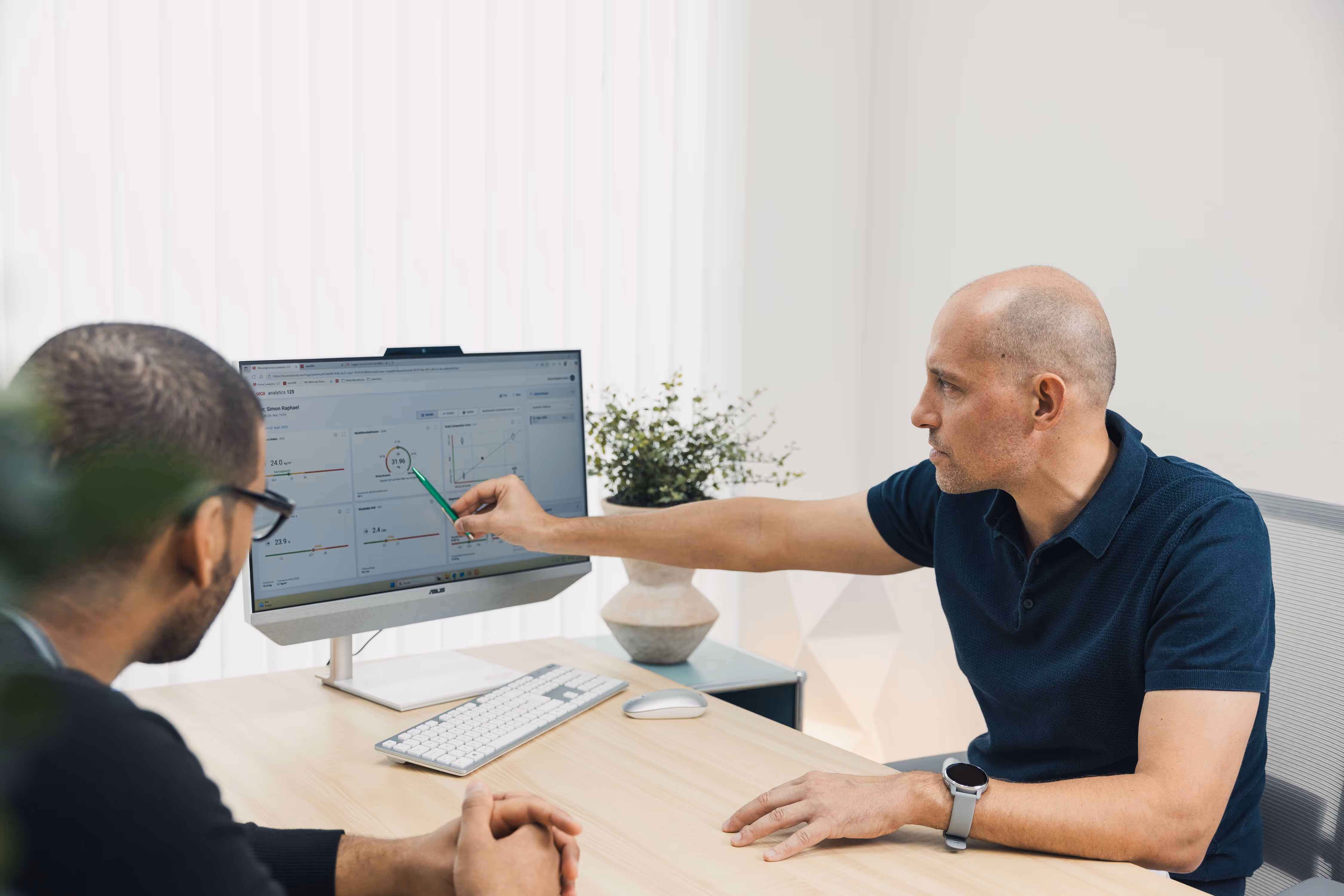Man in navy polo shirt pointing with a pen at data charts on a computer screen while another man watches.