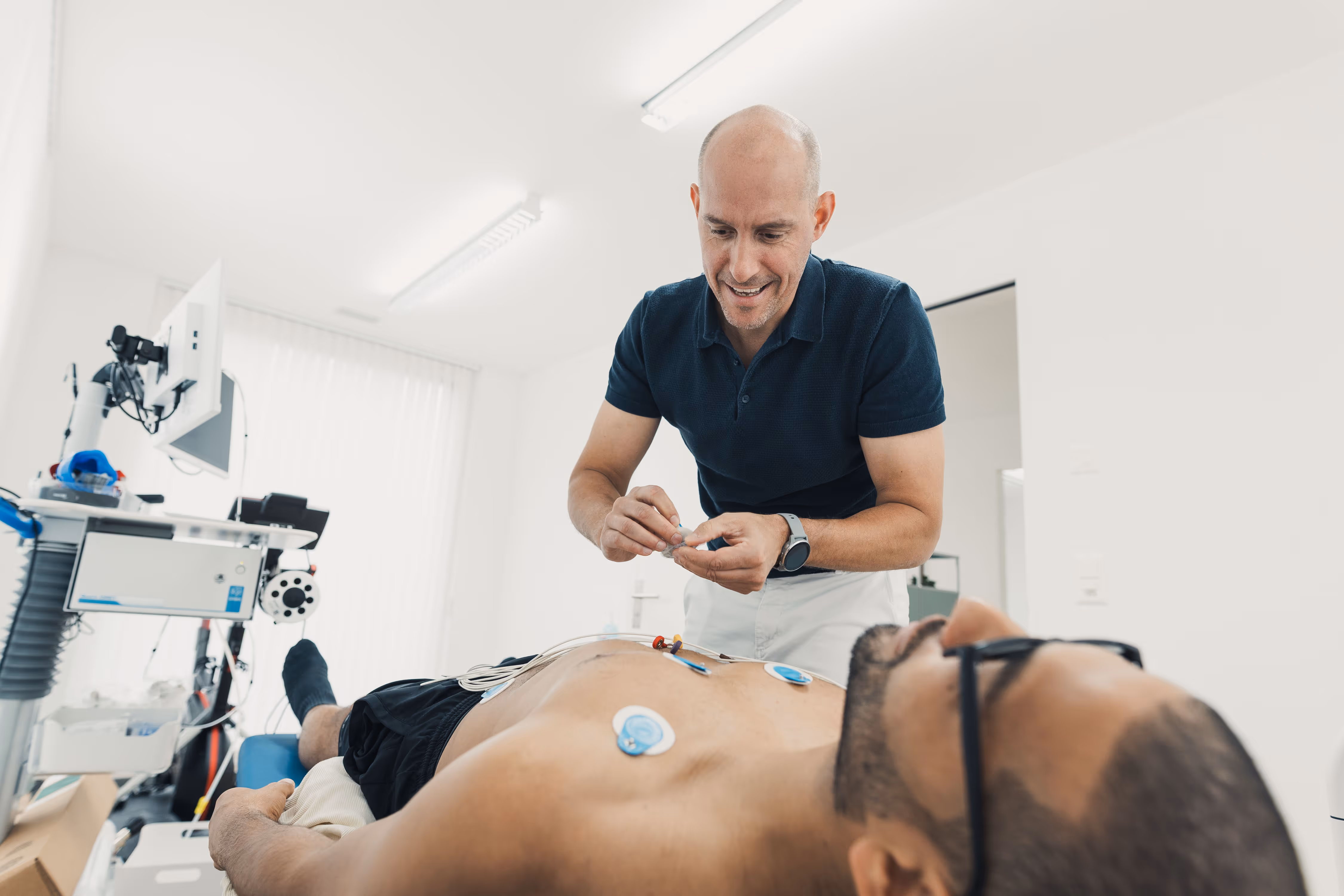 Medical professional attaching electrodes to a shirtless patient lying on an examination table in a clinical setting.