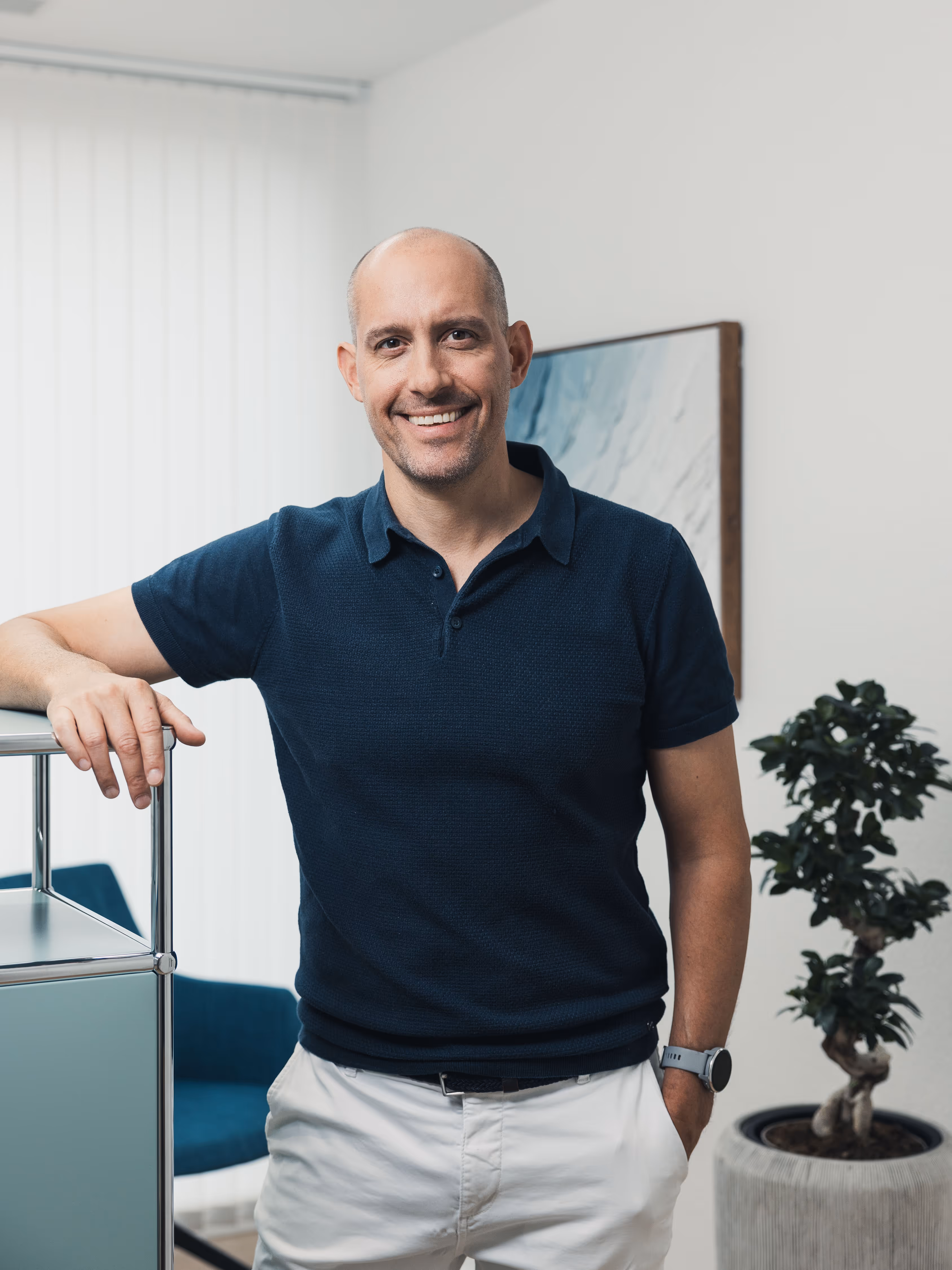 Smiling bald man in a navy blue polo shirt and white pants standing indoors with one hand on a shelf and a plant in the background.