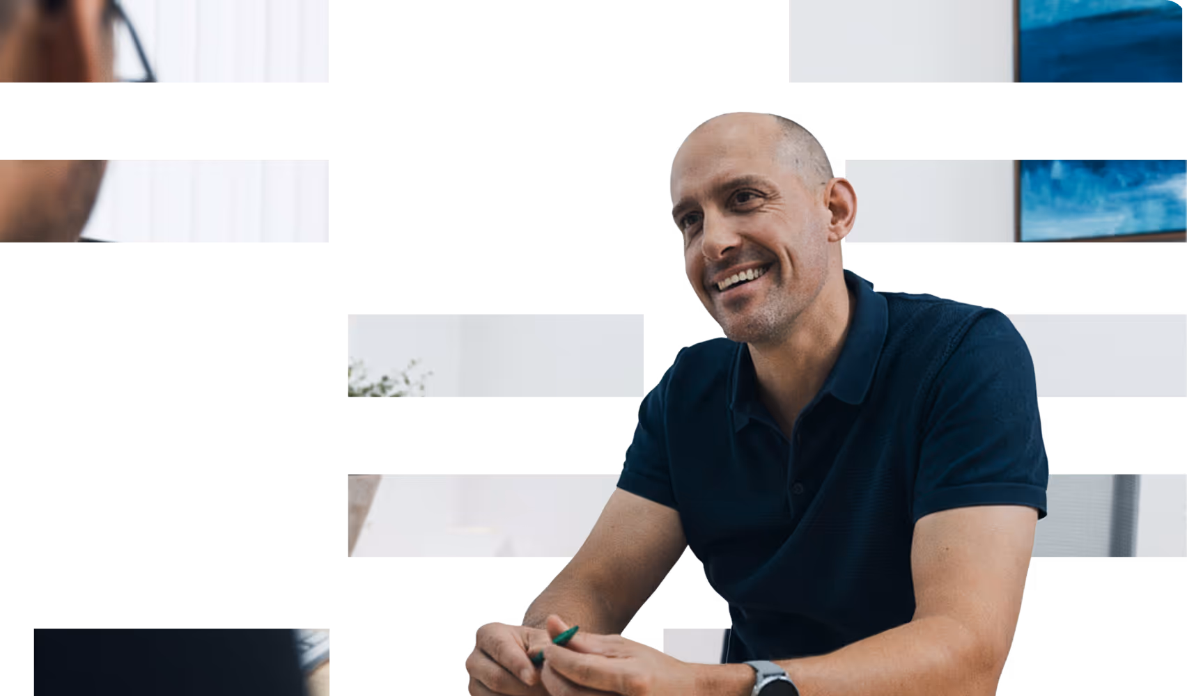 Smiling bald man wearing a dark polo shirt holding a green pen during a conversation in a bright office.